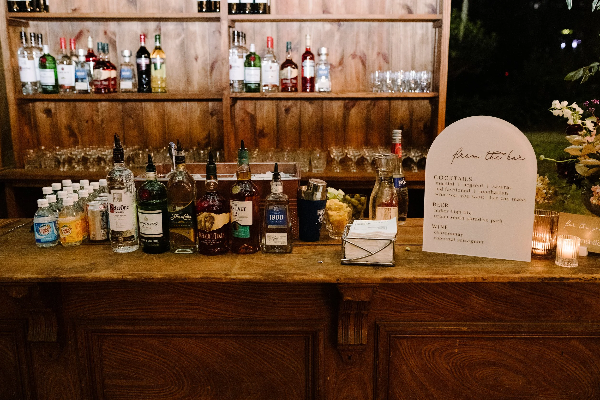 A bar counter with various bottles of alcohol and mixers, a sign listing cocktails and drinks, glassware on the shelves behind, and candles on the counter, with a wooden backdrop and outdoor setting at night.