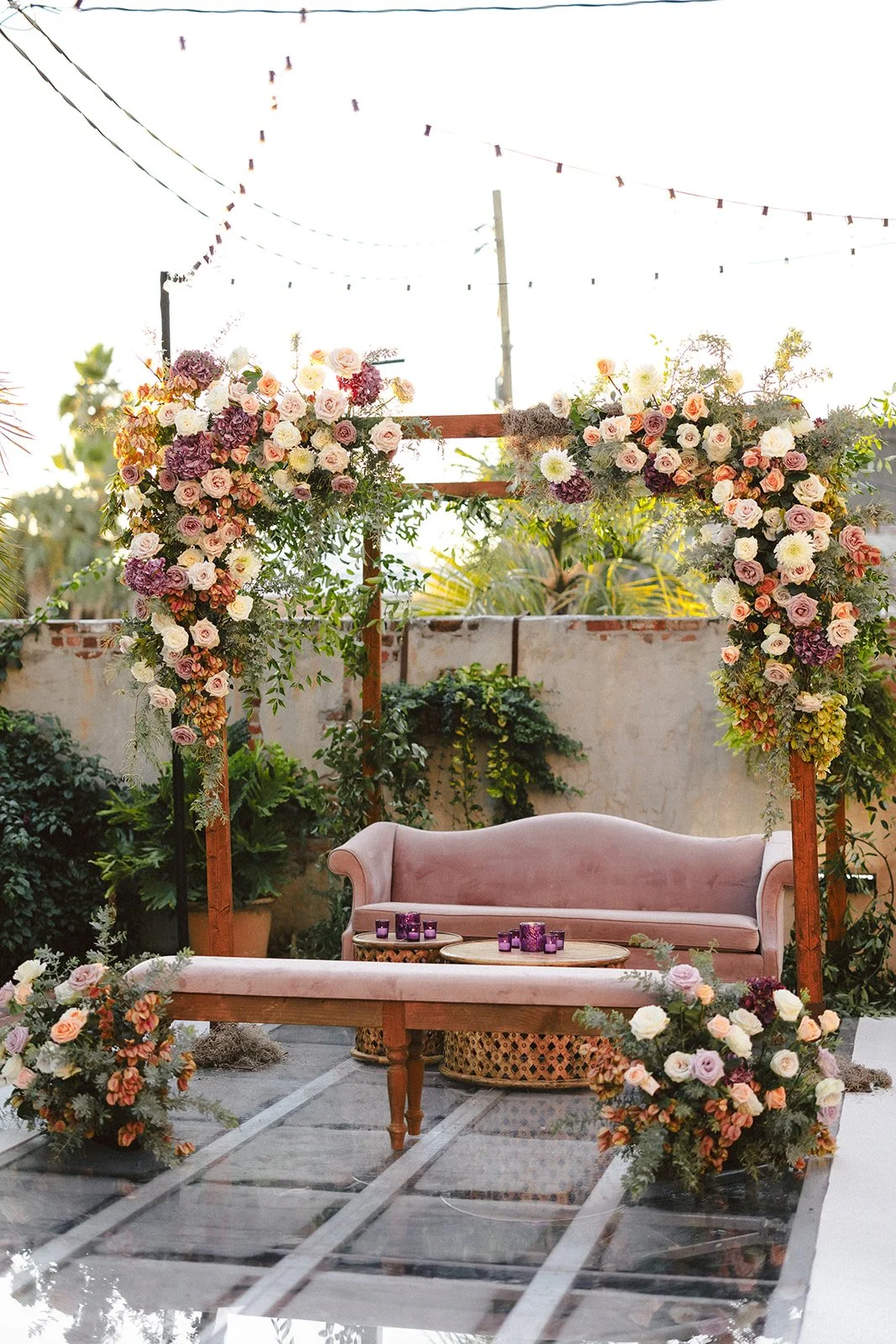A decorated outdoor wedding or event altar with a pink velvet sofa, surrounded by archway of pink, purple, and white flowers with greenery. Purple candles are on small tables, set on a glass floor with a brick wall and string lights overhead.