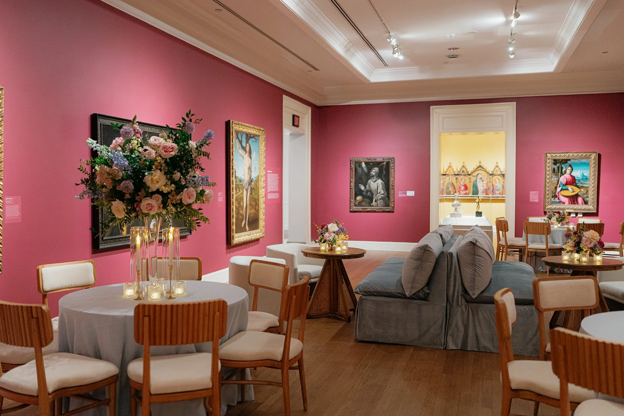A gallery interior with pink walls, floral art, and tables decorated with flowers and candles, featuring seating arrangements including chairs and sofas.