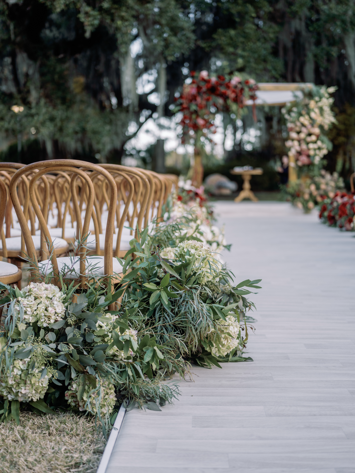 Outdoor wedding ceremony setup with wooden chairs on the left, decorated with lush green foliage and white flowers lining the aisle, leading to an altar with red and pink floral arrangements in a garden setting.