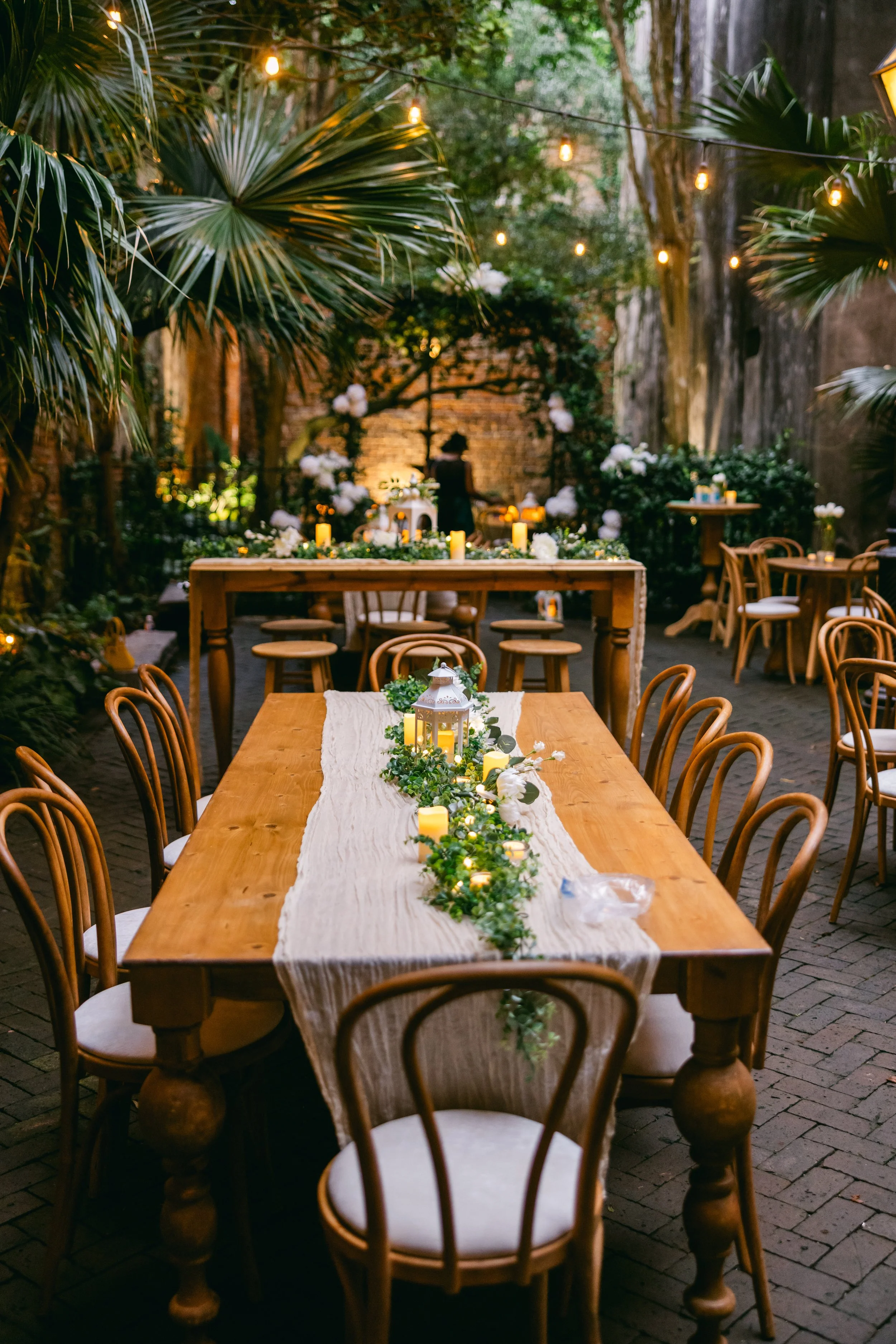 Outdoor seated dinner wedding reception with mixed table seating having a long wooden dining table, dining chairs, pub table and barstools at the New Orleans Pharmacy Museum.