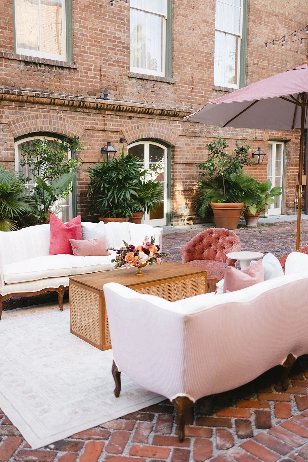 Outdoor patio seating area with vintage style white sofas, pink plush chairs, a wooden coffee table with a floral bouquet, potted plants, an umbrella, and brick walls.