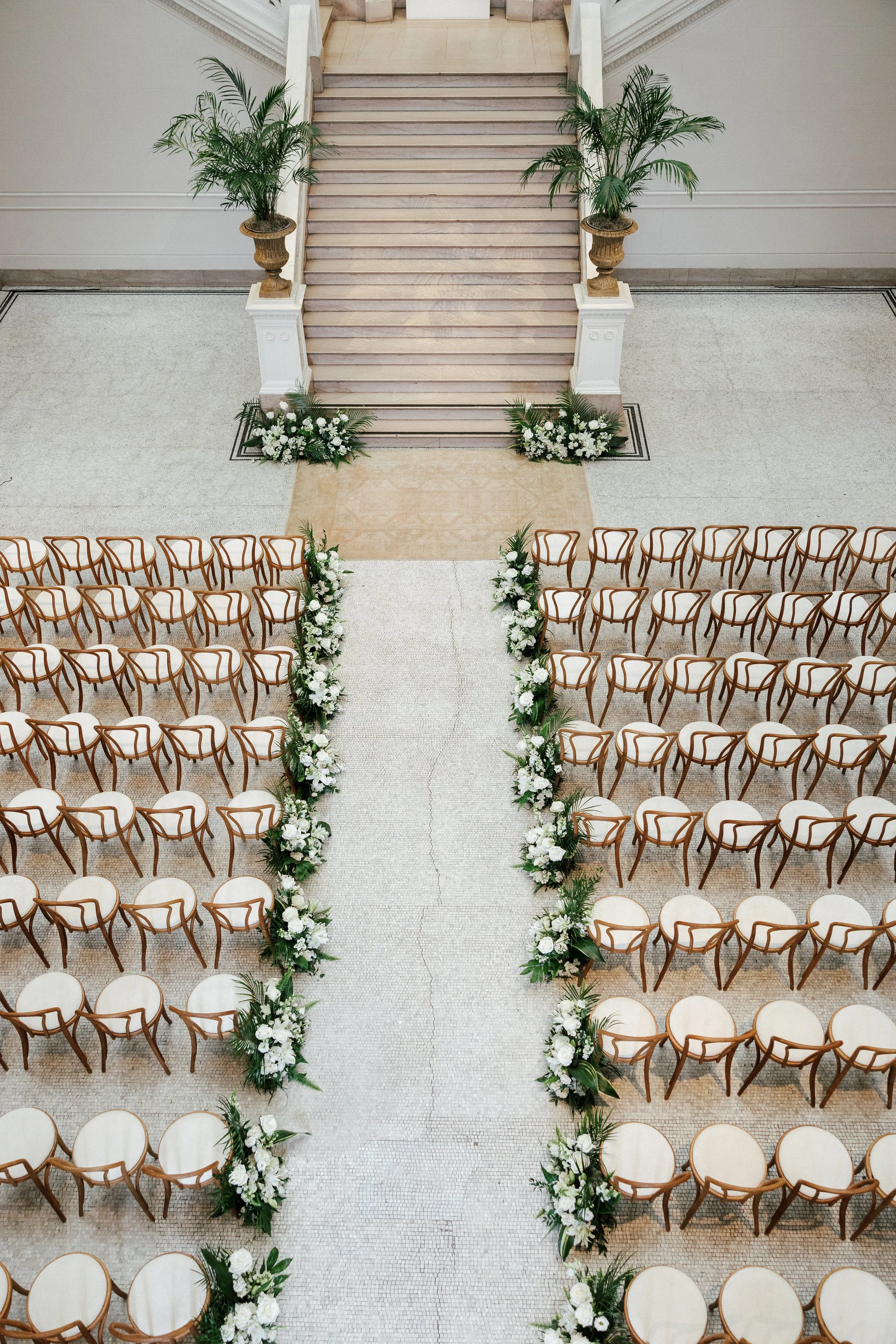 A wedding ceremony setup with cream-colored chairs arranged in rows on each side of an aisle decorated with white flowers and greenery, leading to a stairway with potted plants on each side.