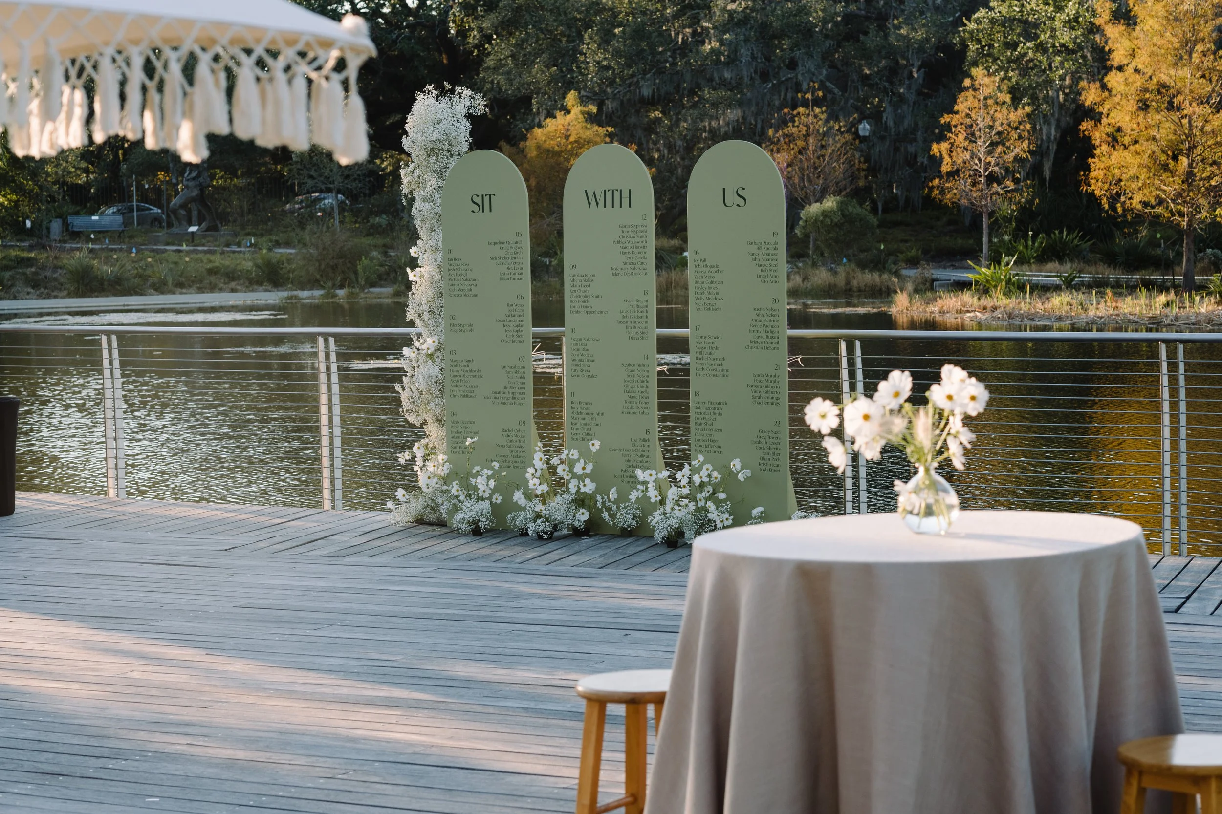 A lakeside outdoor wedding setup with a floral display and a board with seating arrangements, surrounded by trees with fall foliage.