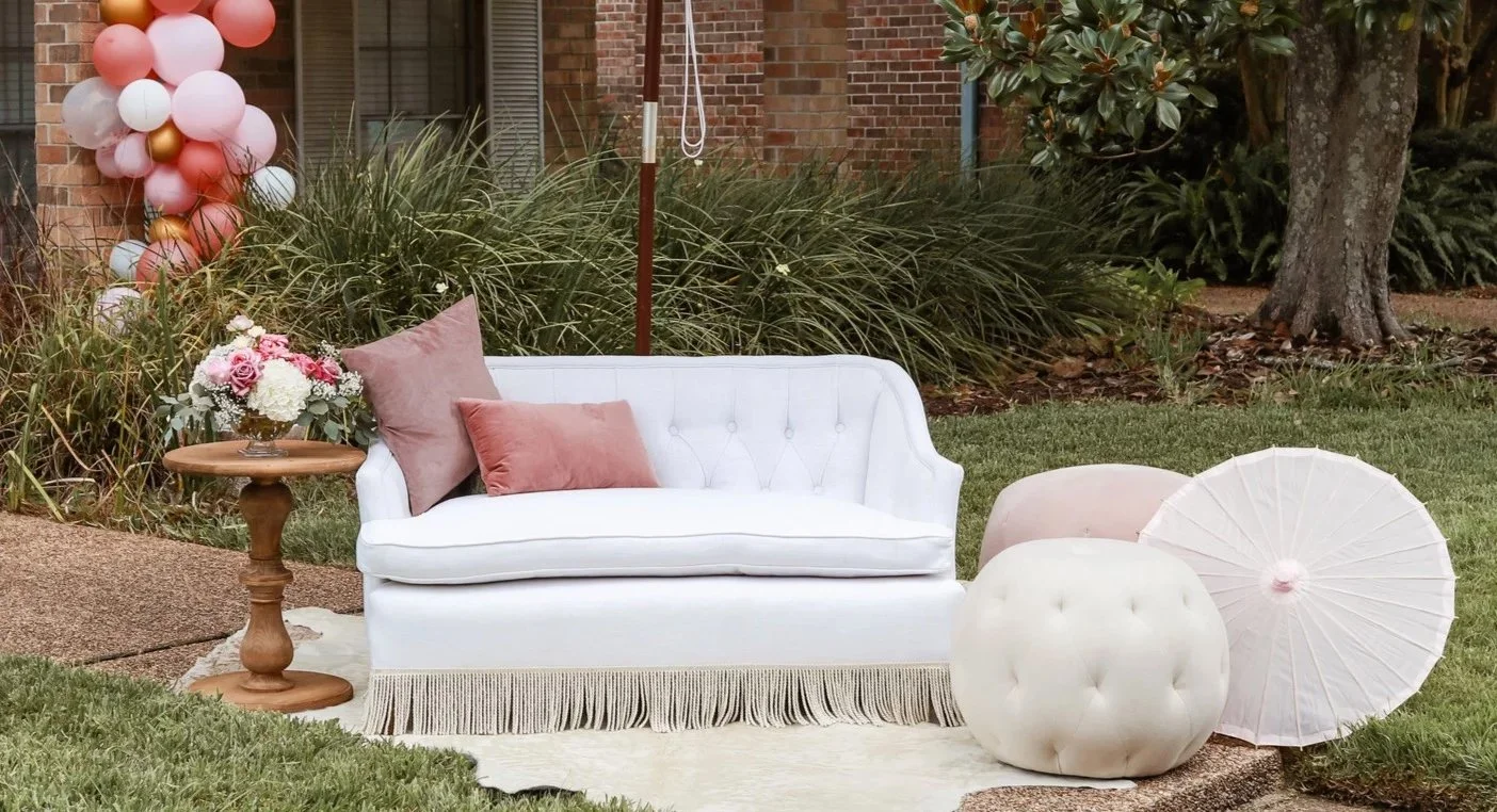Outdoor scene with a white vintage loveseat, pink and beige pillows, a small round wooden table with a flower arrangement, pink and white balloons, a white umbrella, a tufted ottoman, and a garden with trees and bushes.