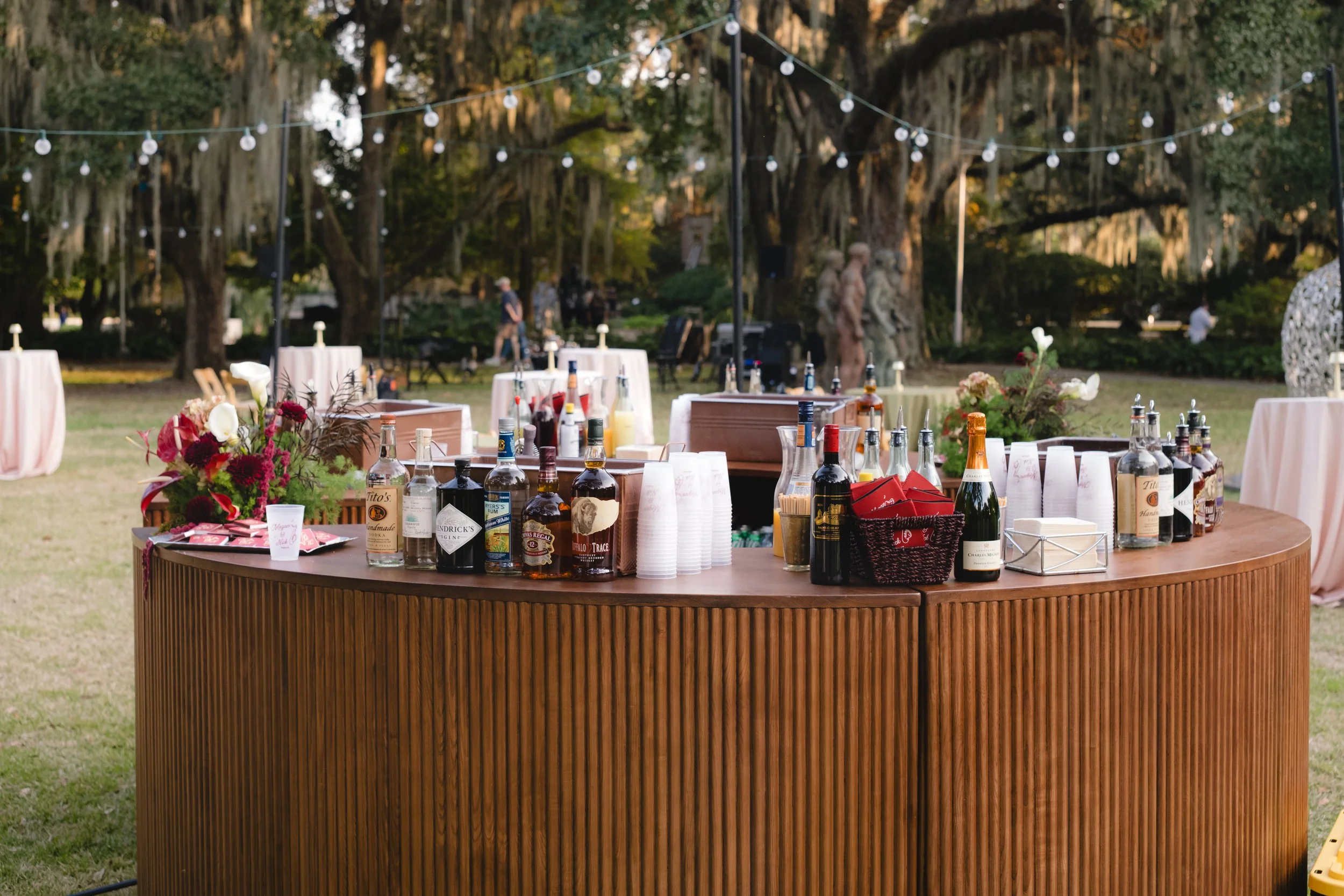 Outdoor event setup featuring a curved wooden bar counter with various bottles of alcohol, mixers, cups, and a floral arrangement, set against a background of large trees and string lights.