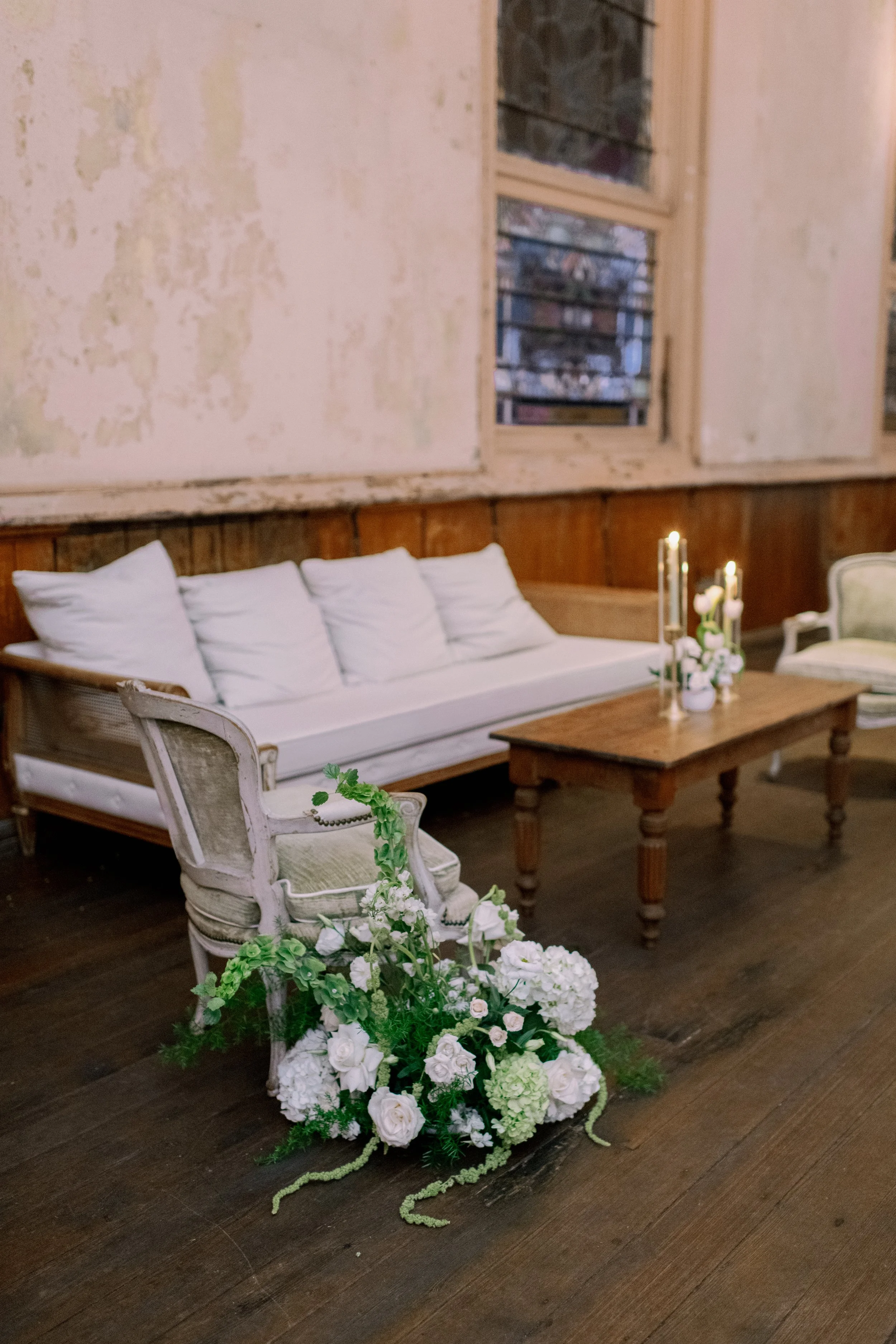 A room with cream-colored, slightly distressed walls, a large window with multiple panes, a white vintage sofa with multiple pillows behind a wooden coffee table, white and green floral arrangements in a basket on the floor, and a candle holder with 
