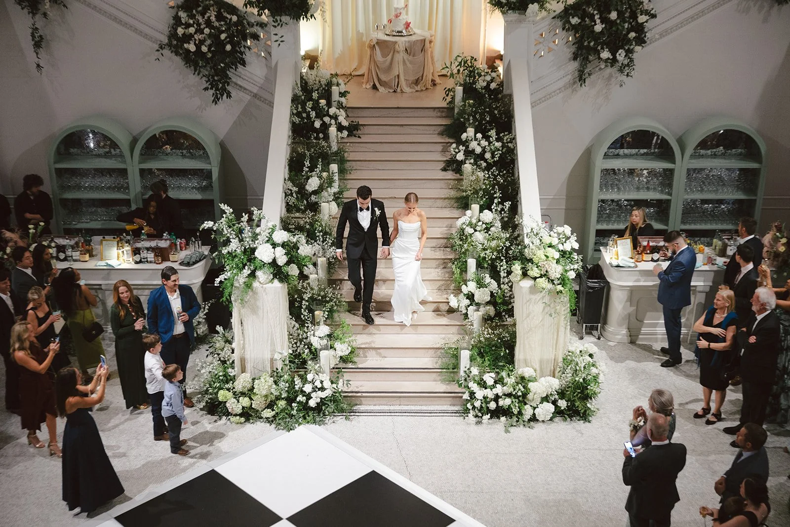 A wedding reception with a bride and groom descending a floral decorated staircase. The bride is in a white strapless gown, and the groom is in a black tuxedo. Guests are watching and taking photos, with some at a bar area to the sides.