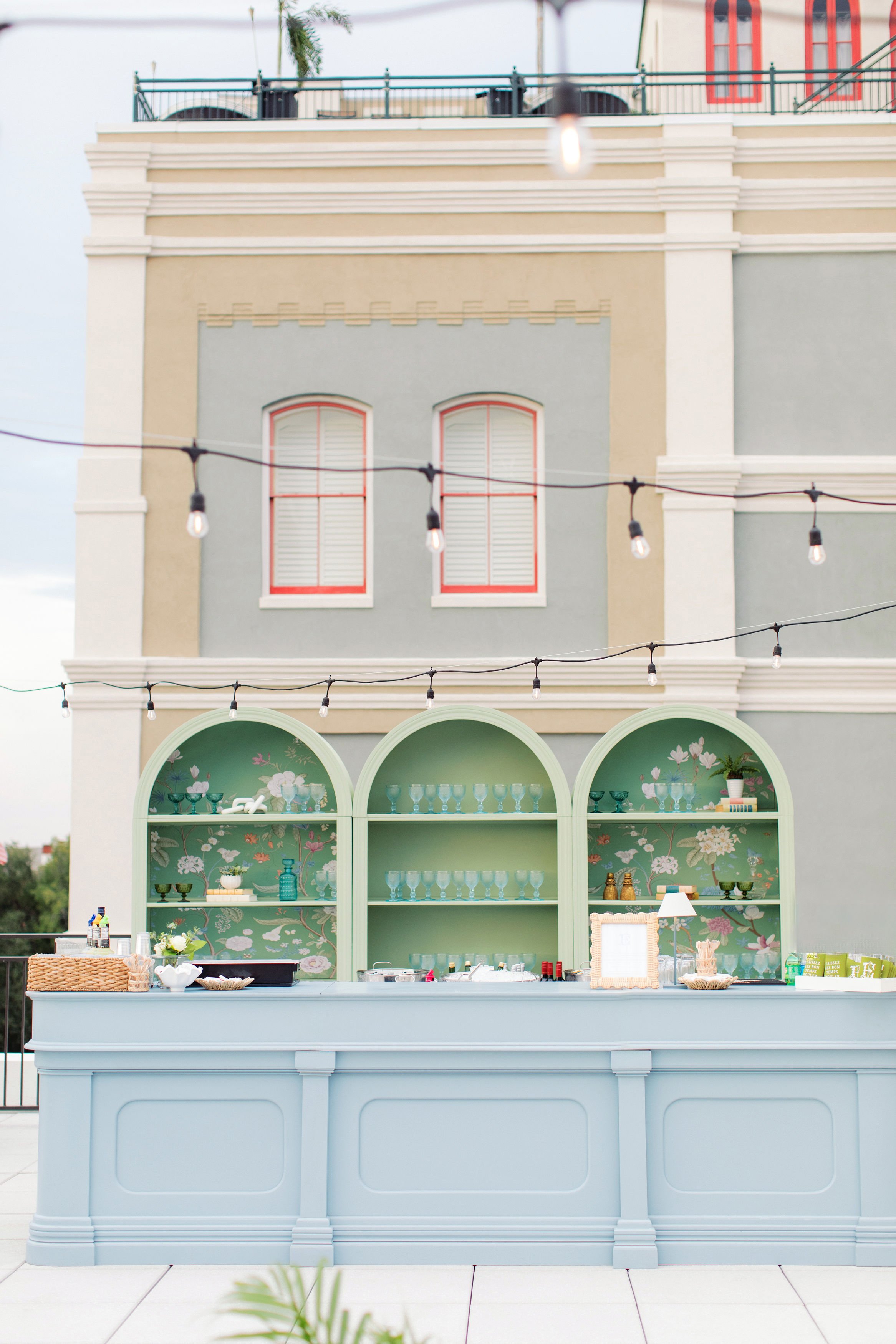 An outdoor display of glassware and decor on pastel green shelves and furniture against a building with pastel yellow, beige, and green walls, and string lights hanging overhead.