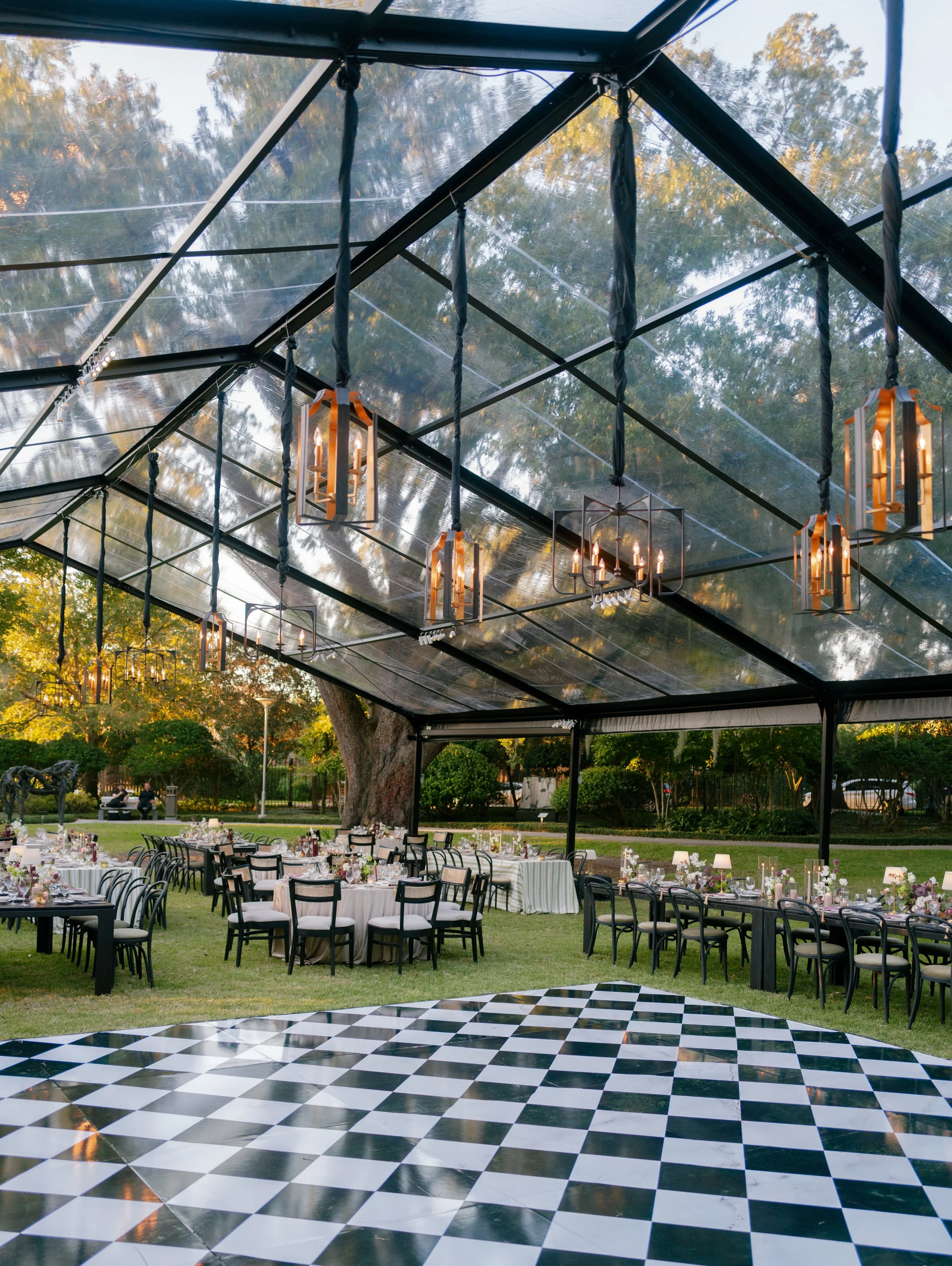 Outdoor event setup in a garden with round tables, black chairs, and floral centerpieces, covered by a glass canopy with hanging chandeliers, trees, and a checkered dance floor visible in the foreground.
