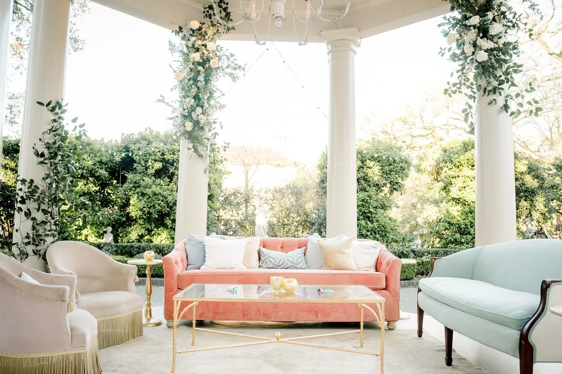 Sunlit outdoor patio with pastel-colored sofas, white pillars, and lush green foliage.