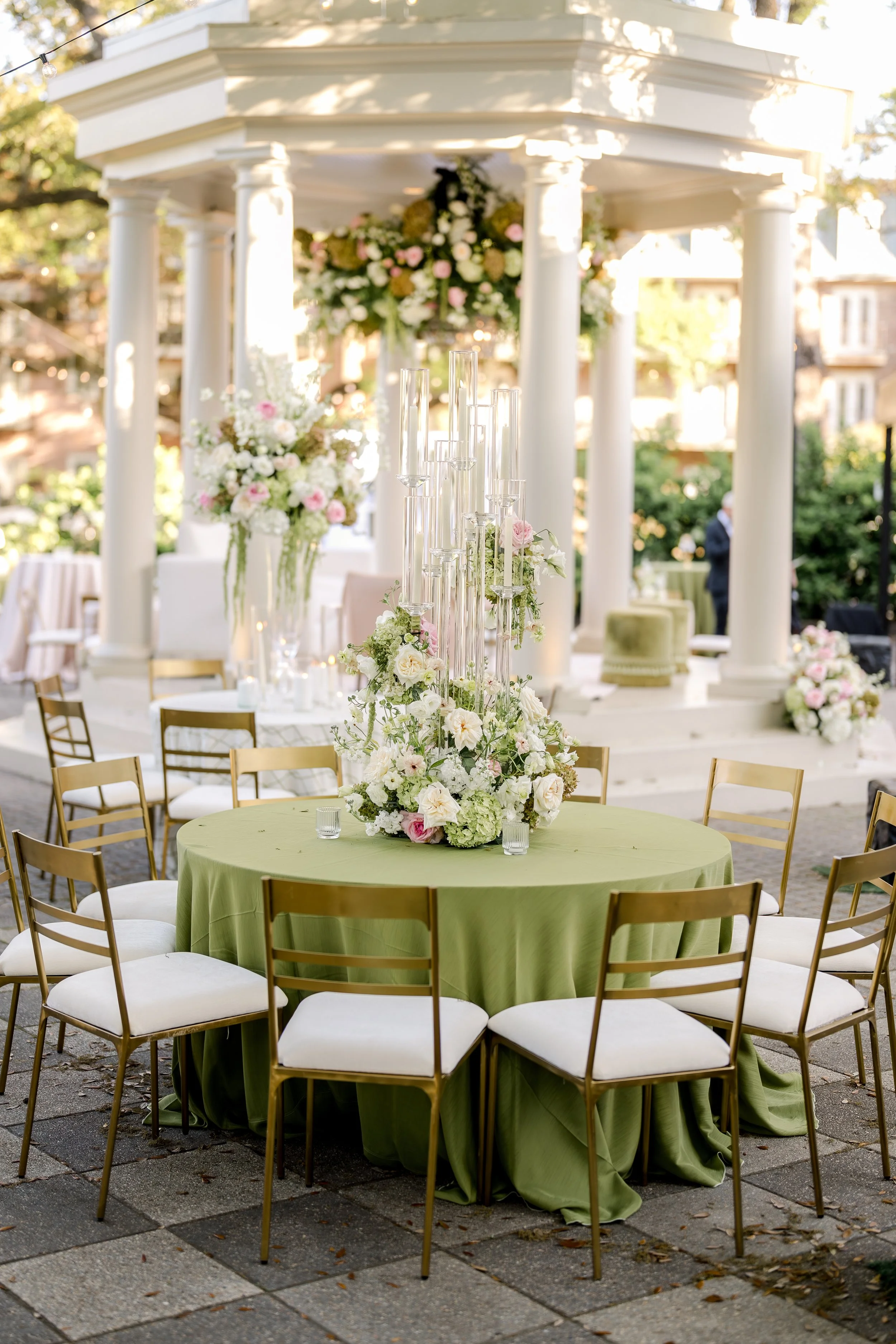 A round table with a green tablecloth and a floral centerpiece surrounded by gold chairs with white cushions. In the background, there is a white pavilion with columns and hanging floral arrangements, decorated for an outdoor event.