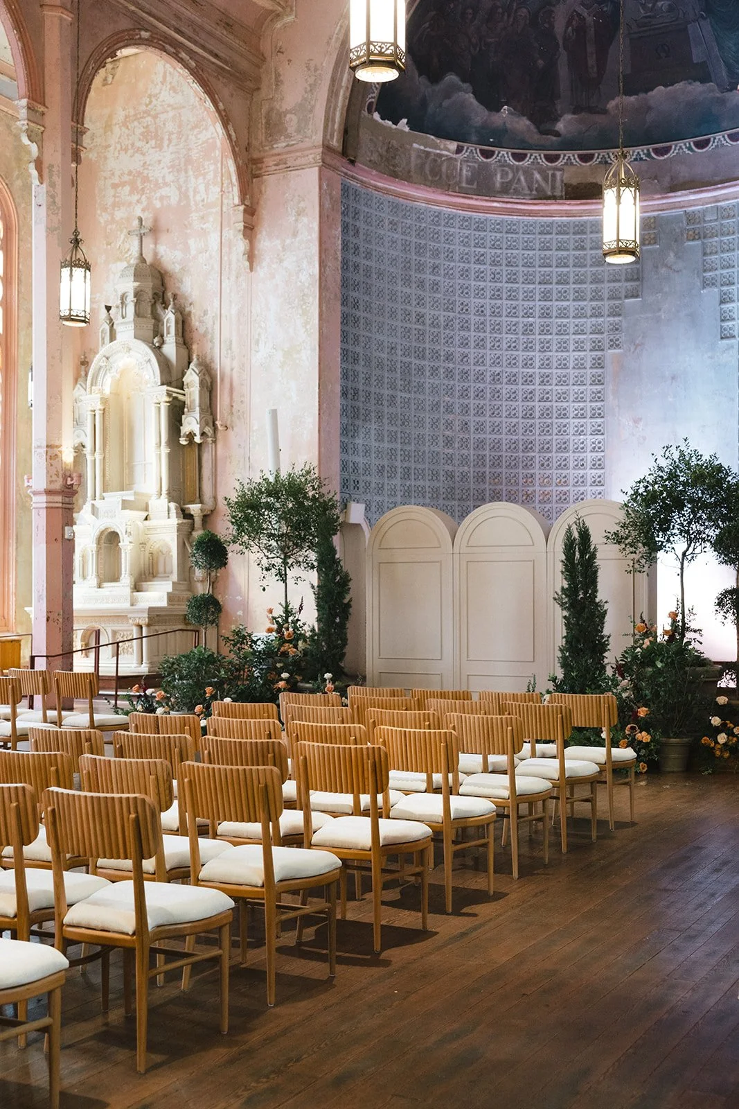 Interior of a church or chapel decorated for a wedding, with rows of wooden chairs with white cushions, greenery and flowers, an ornate white altar, and hanging lanterns.