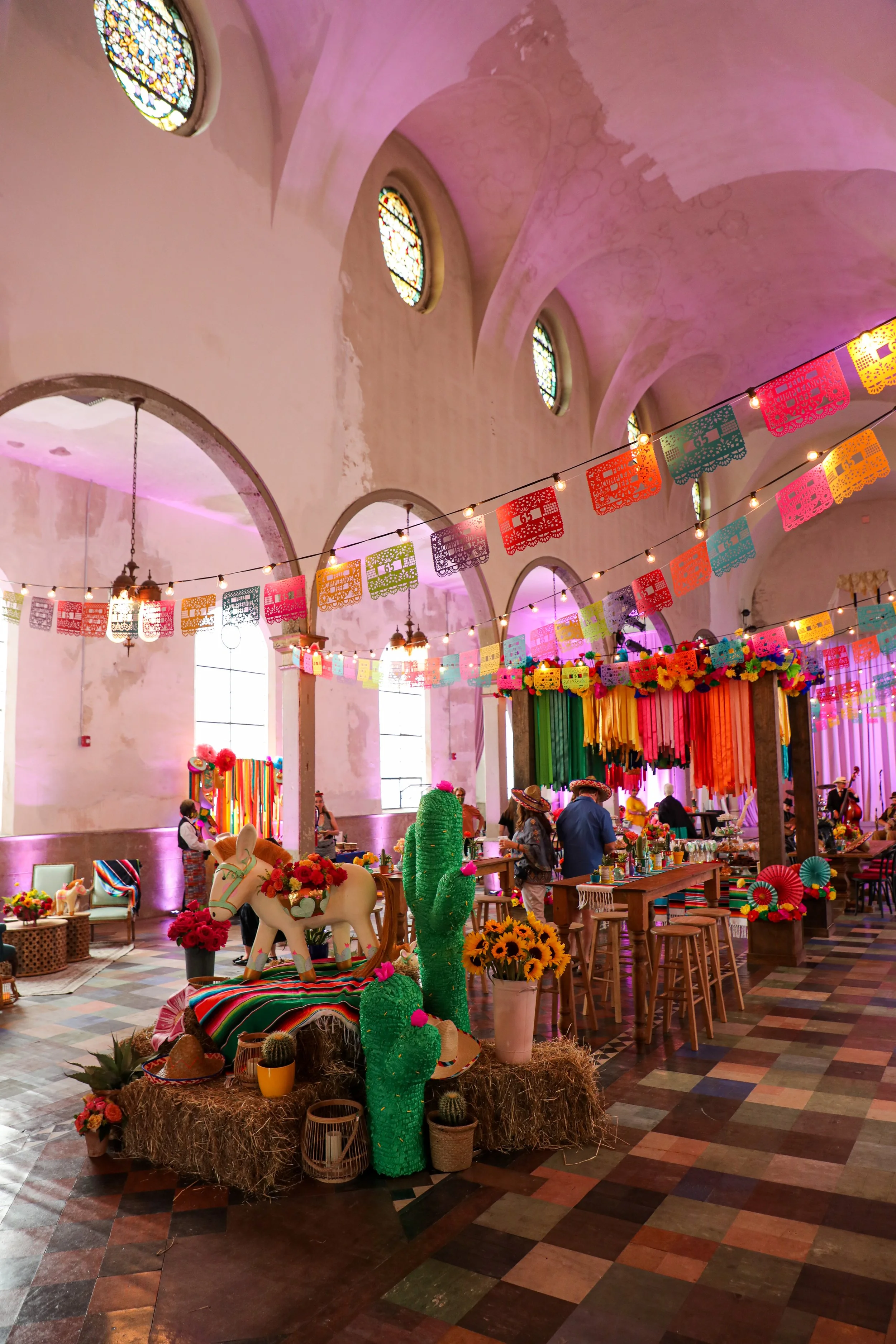 Interior of a large church decorated with colorful papel picado banners, flowers, and cactus figures for a festive celebration. The church has stained glass windows, high arched ceilings, and people wearing sombreros.