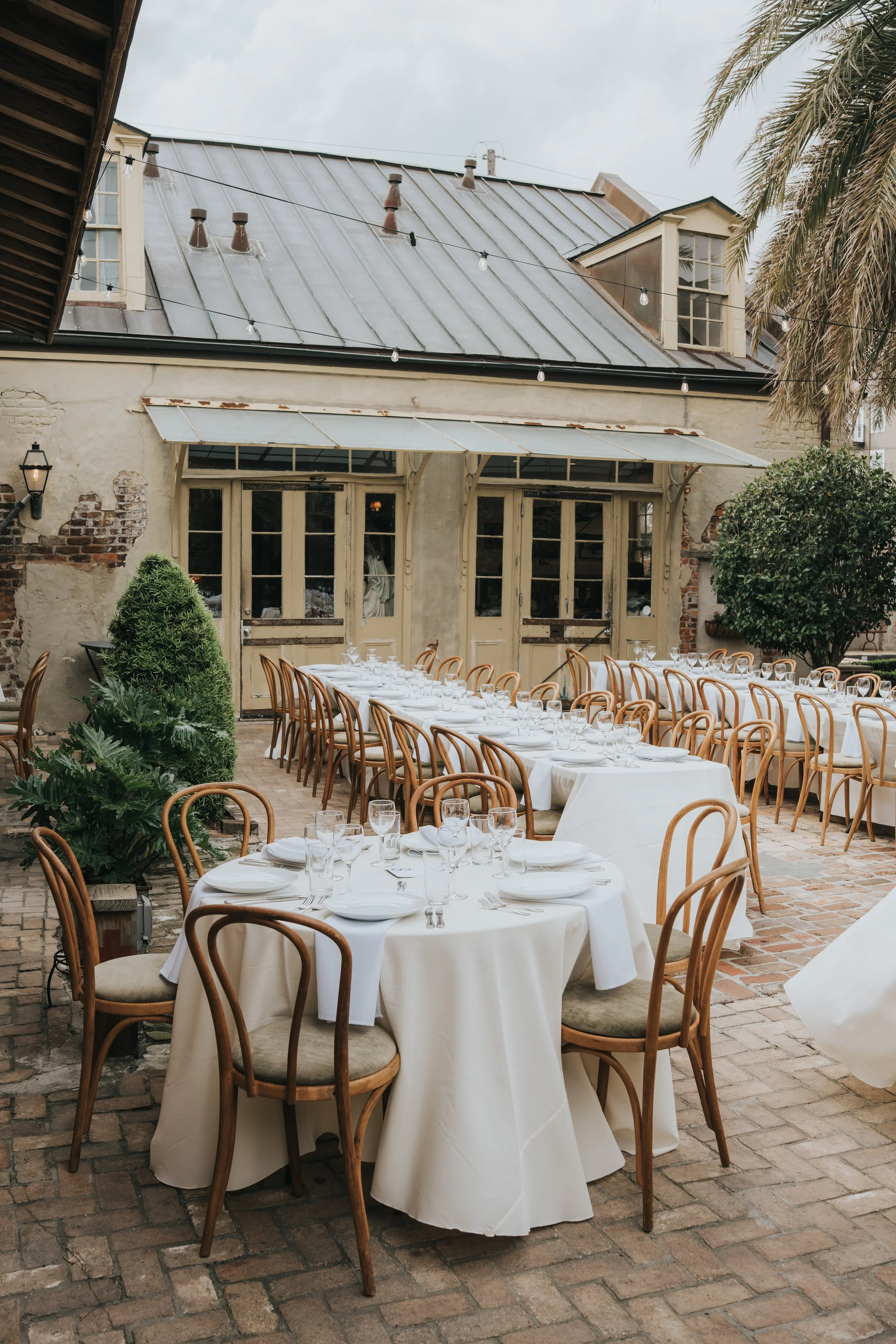 Outdoor dining setup with round tables covered in white tablecloths, surrounded by wooden chairs with beige cushions, set with glasses, silverware, and plates in front of a rustic building with large windows and a metal roof. String lights are hung a
