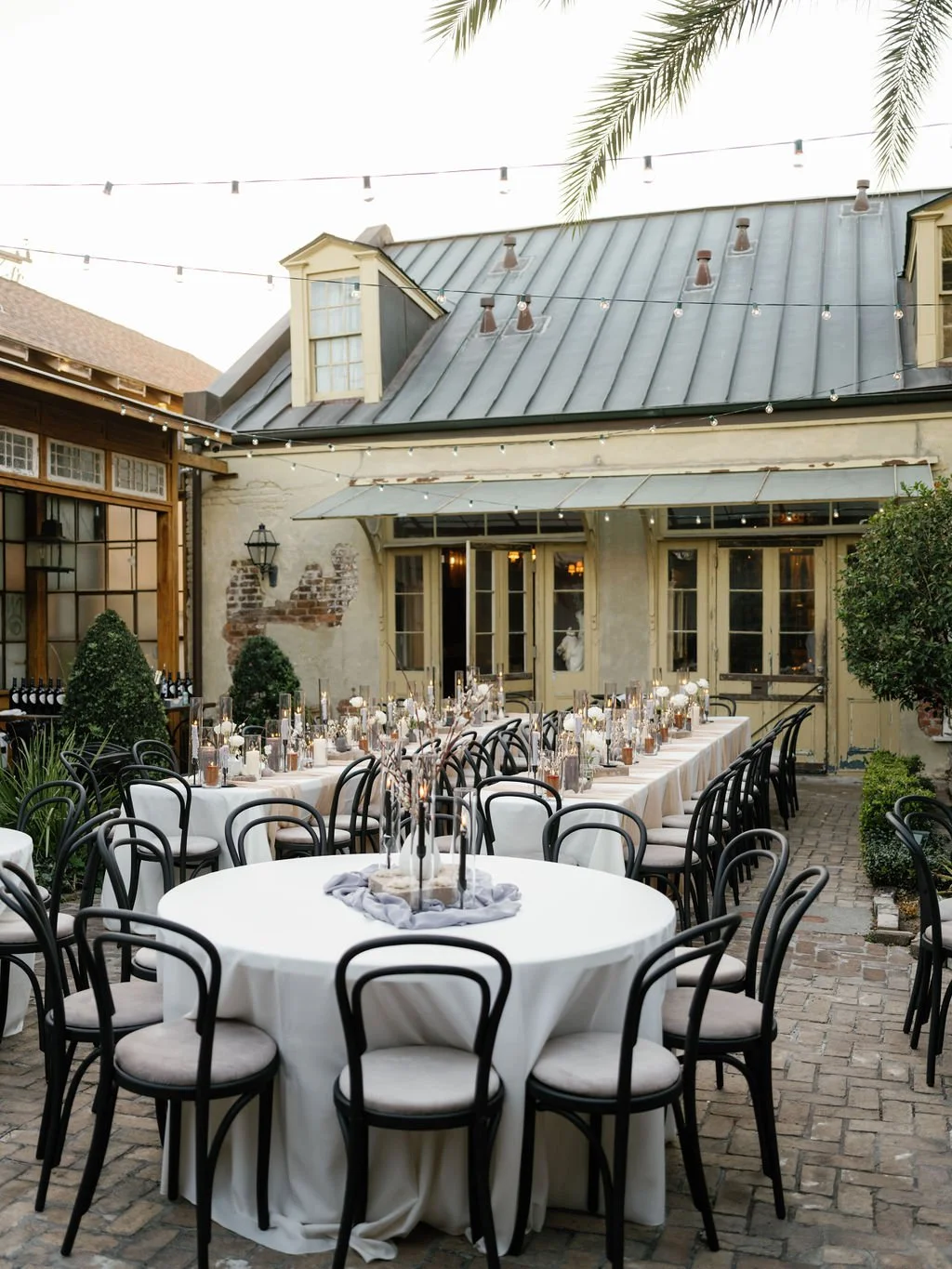 Outdoor wedding reception setup with round tables covered in white tablecloths, adorned with candles and floral arrangements, situated in a courtyard with a brick patio, surrounded by greenery and an old building with string lights hanging overhead.