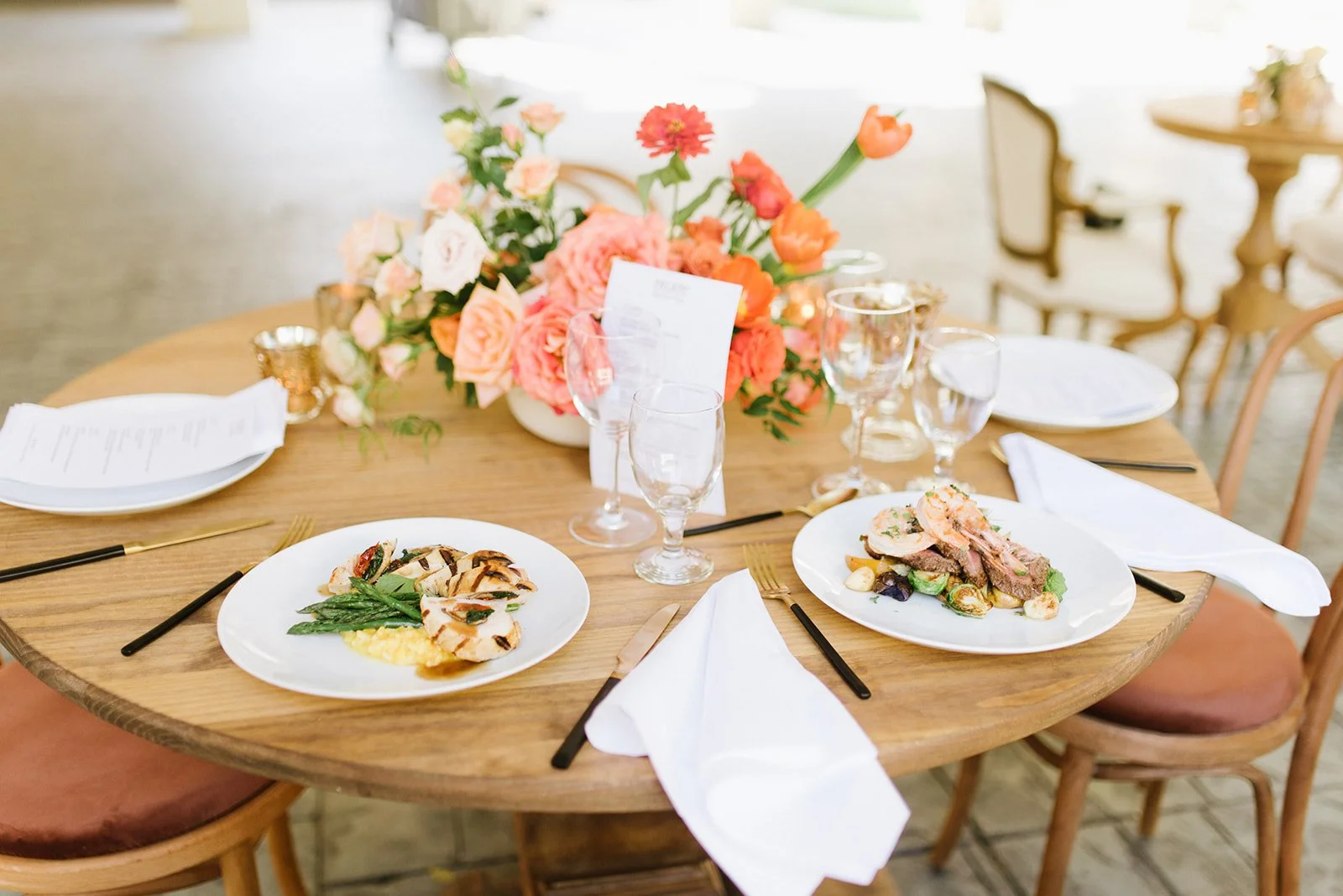 A round wooden dining table set for a formal meal with plates of food, wine glasses, and silverware. A large floral centerpiece with pink and orange flowers decorates the table. Empty chairs surround the table within a dining room.