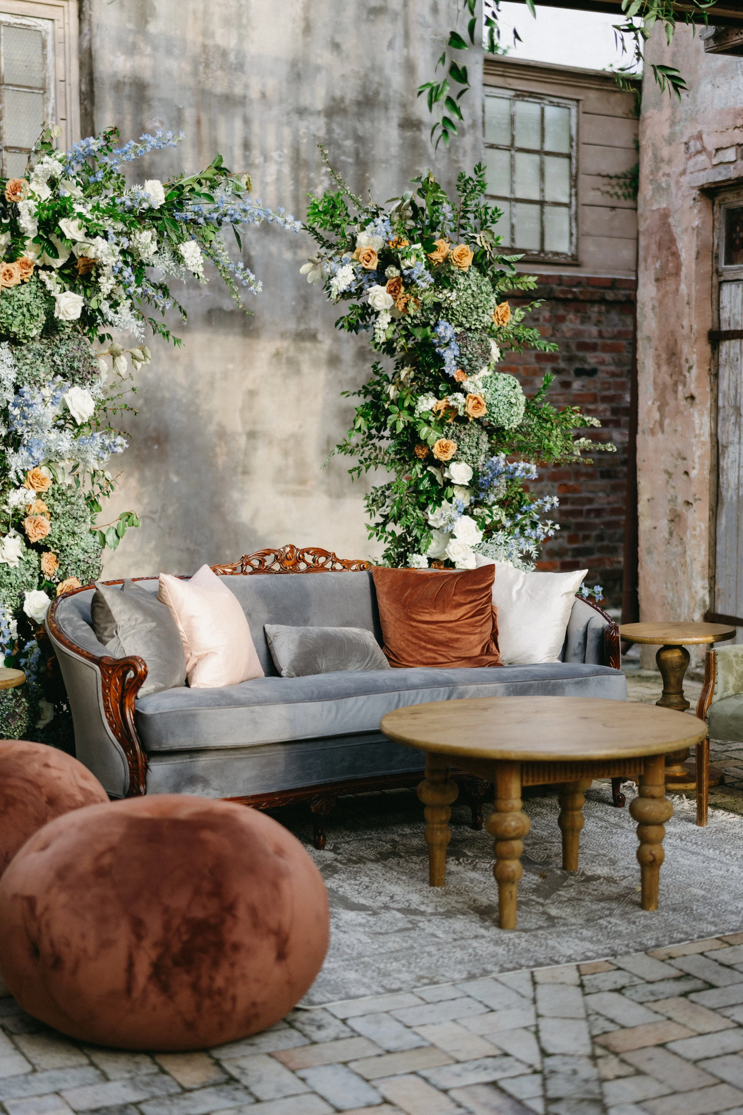 A vintage gray sofa with decorative pillows in various pastel colors, situated in front of a large floral arrangement with white, blue, and peach flowers, set in an industrial-style outdoor space with exposed brick and concrete walls, a small wooden 