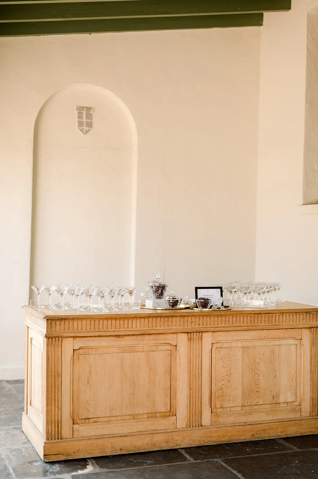 A wooden sideboard with empty glassware and bowls with chocolates, situated against a cream-colored wall with a small, arched decorative inset.
