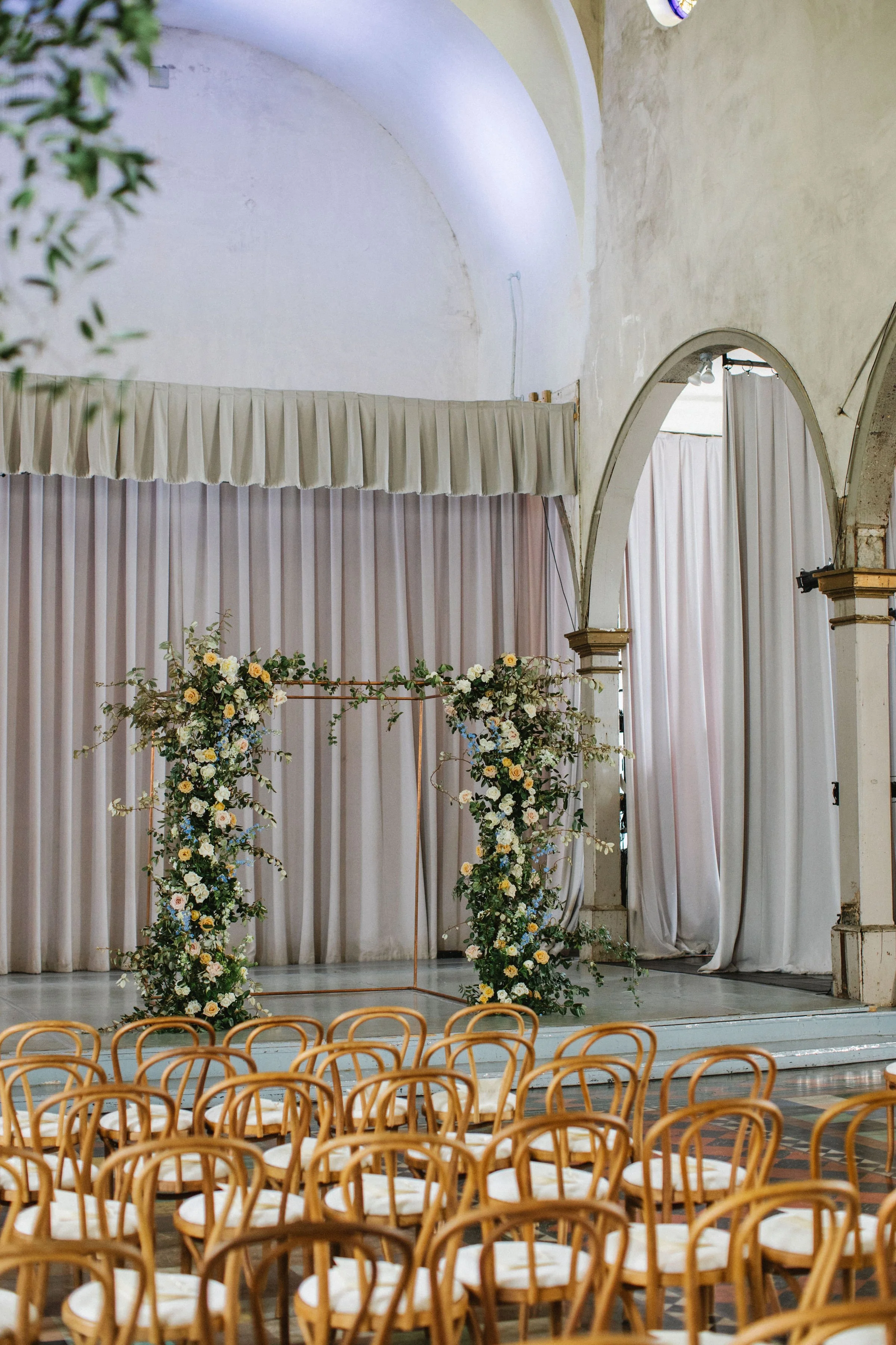 Wedding ceremony setup with rows of wooden chairs facing a floral arch and a backdrop of beige curtains inside a building with high ceilings and arched doorways.