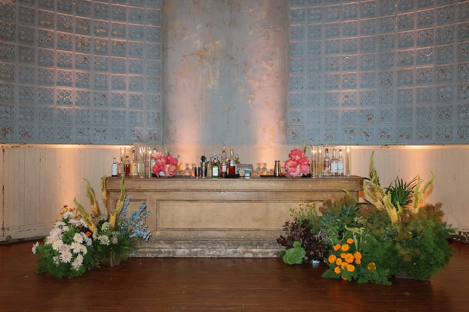 A vintage wooden bar with flowers and bottles, set against a weathered pastel blue wall with floral-patterned tiles in a rustic, elegant setting.