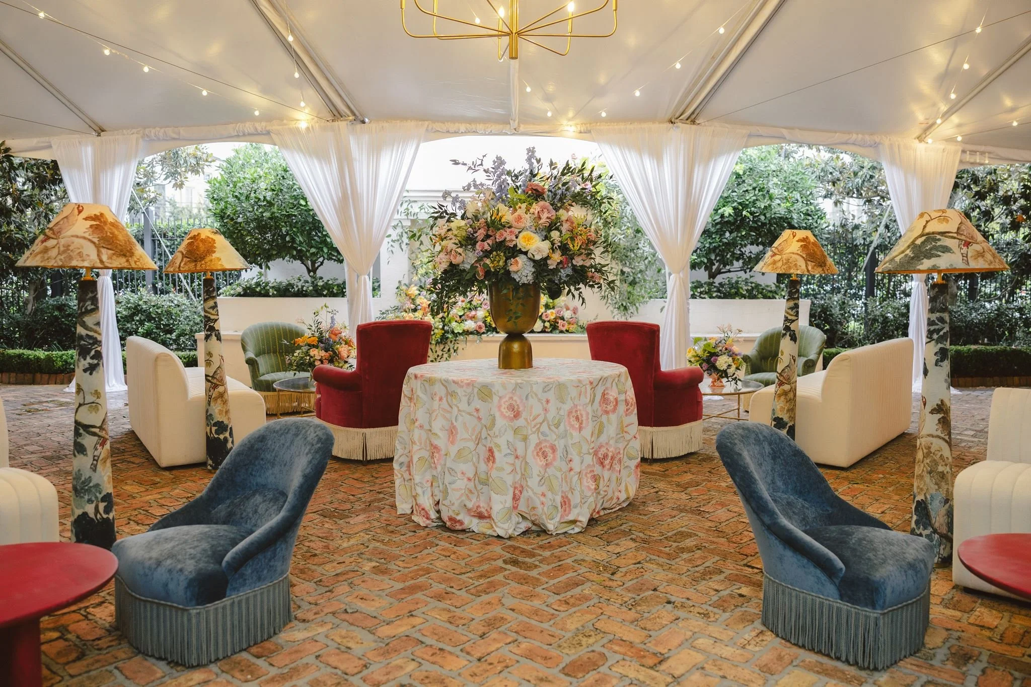 Indoor event space with a large floral arrangement on a round table, surrounded by velvet chairs, with lamps, string lights, and greenery outside visible through windows with white curtains.
