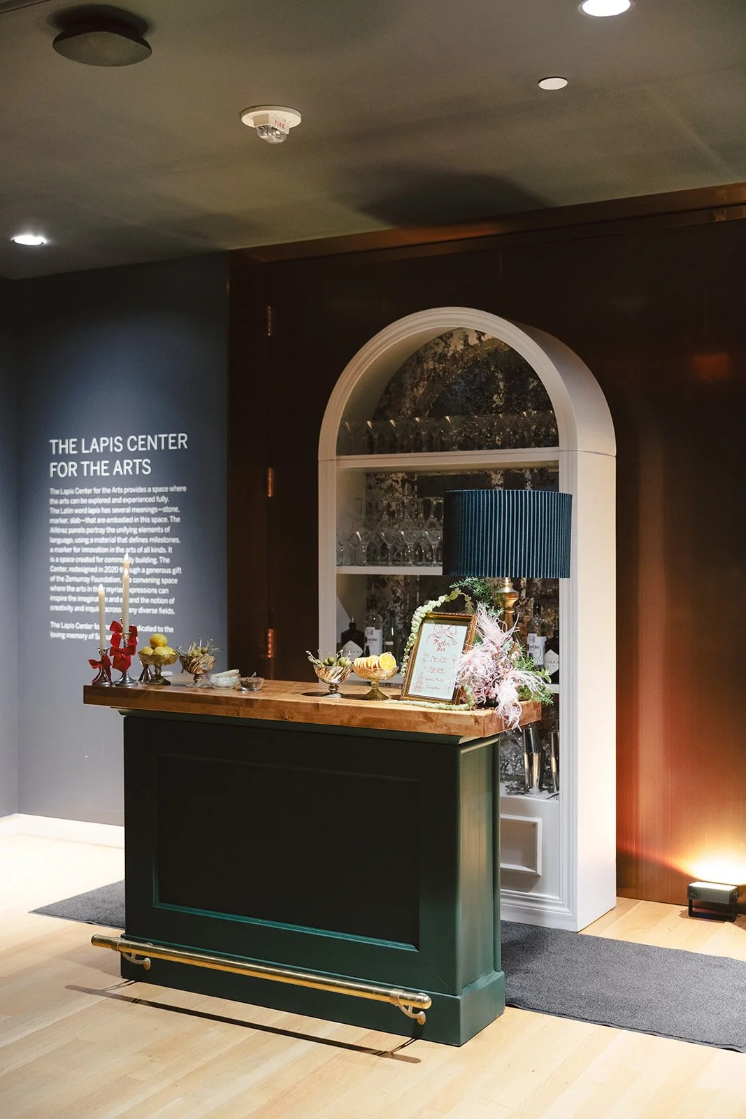 A reception counter at The Lapis Center for the Arts with decorative items and a menu, behind a dark arch and a wall with informational text about the space.