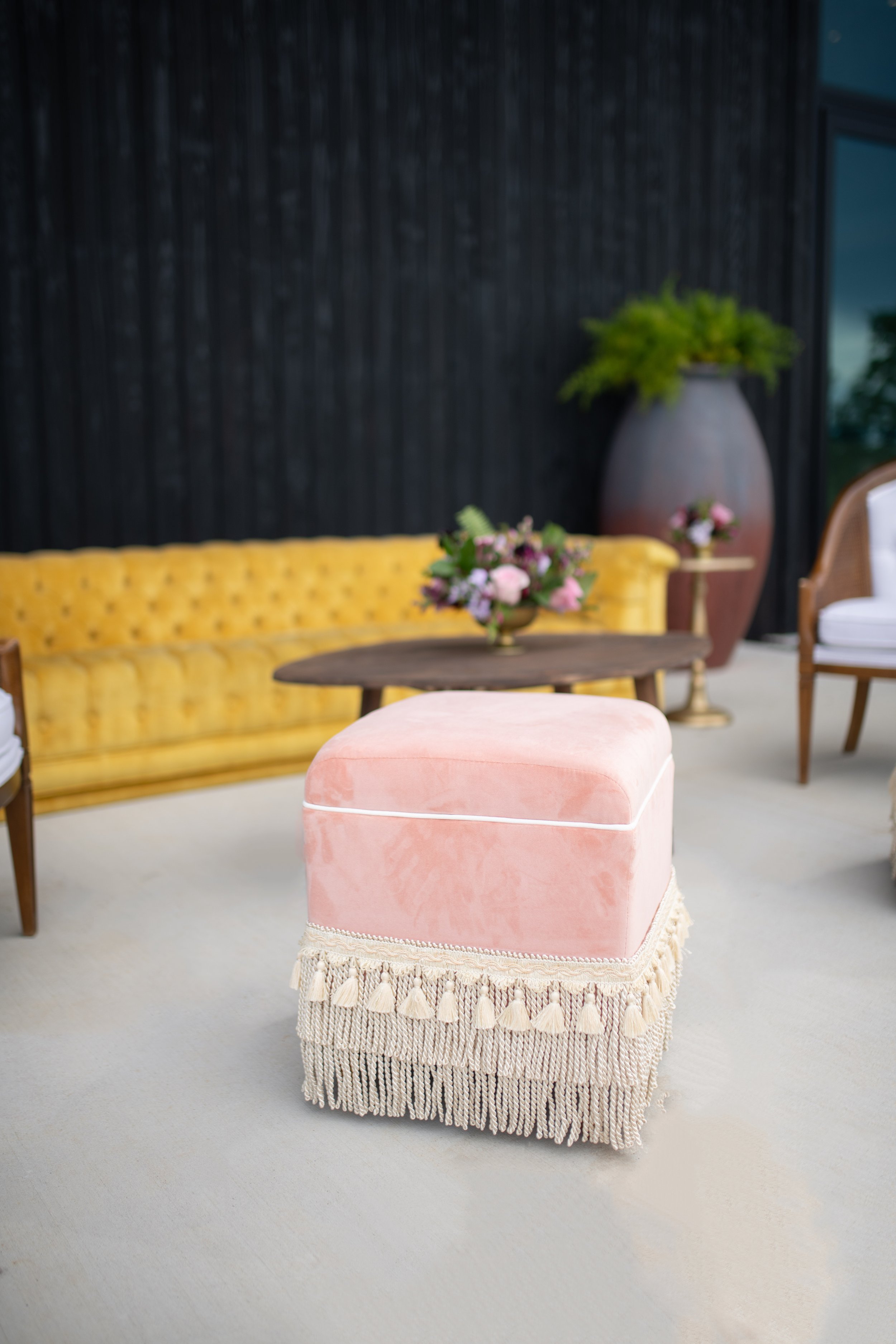A pink upholstered ottoman with a fringed base sits on a light-colored carpet in a room with a yellow tufted sofa, a dark wooden round coffee table with a flower arrangement, and various chairs and large plant pots in the background.
