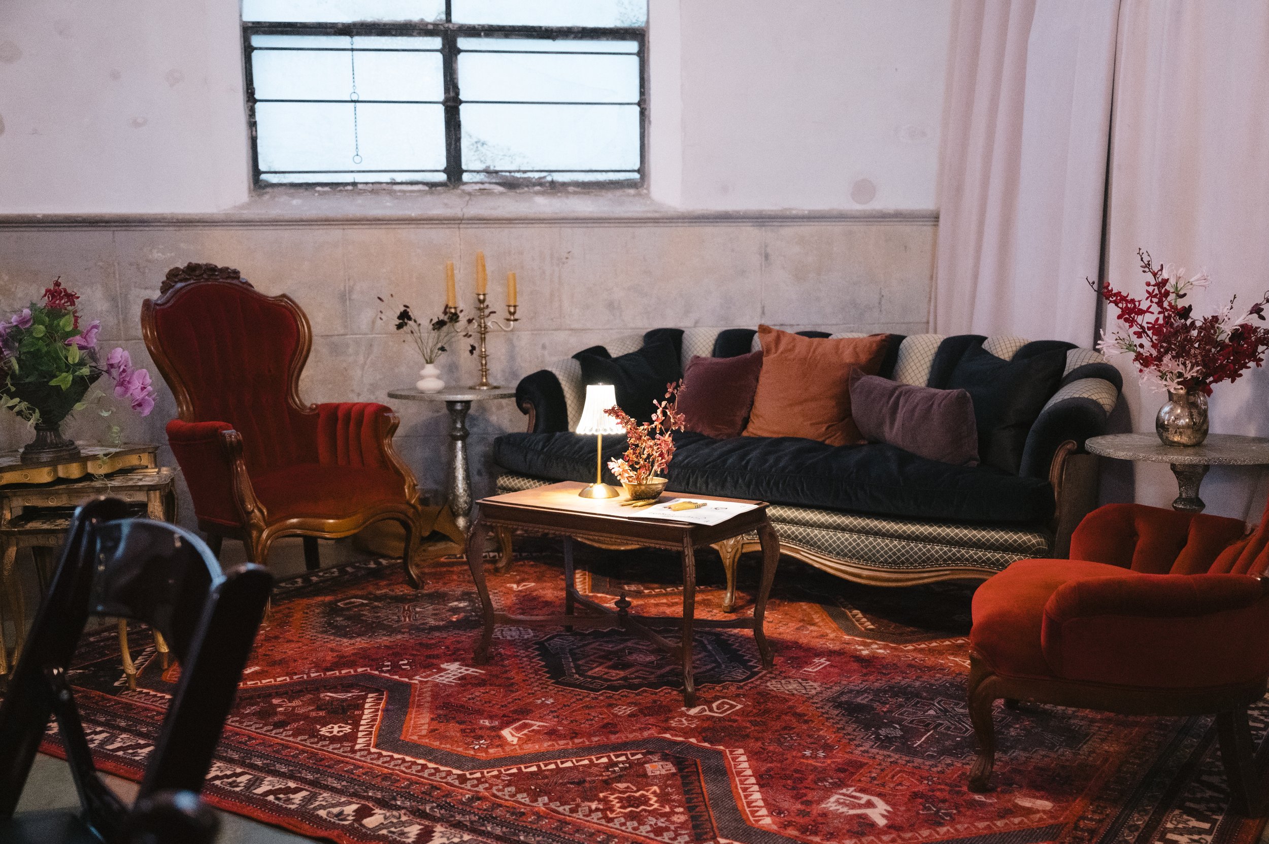 Interior of a living room with vintage furniture, including a red velvet armchair, a black and checkered sofa with multiple pillows, and various decor items like a lamp, a candelabra, and flower arrangements. The room has a large window with metal ba