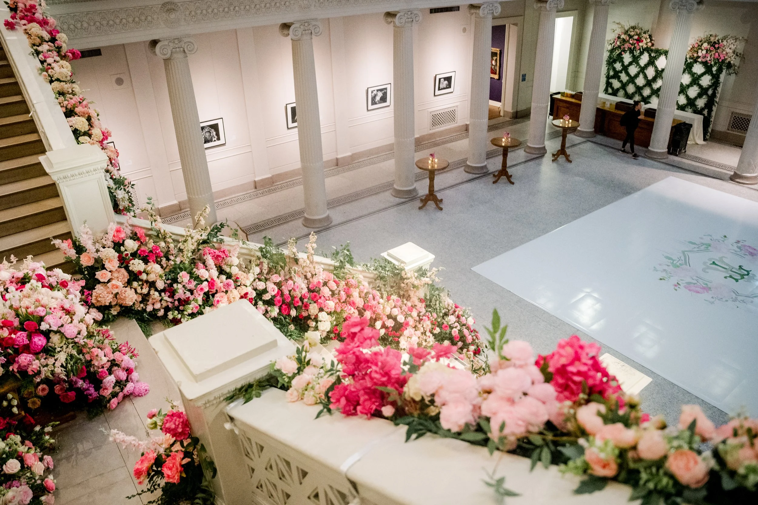 Indoor area decorated with a large floral arrangement of pink and white flowers, columns, framed pictures on the wall, a small table with a candle, and a person standing near a reception desk.