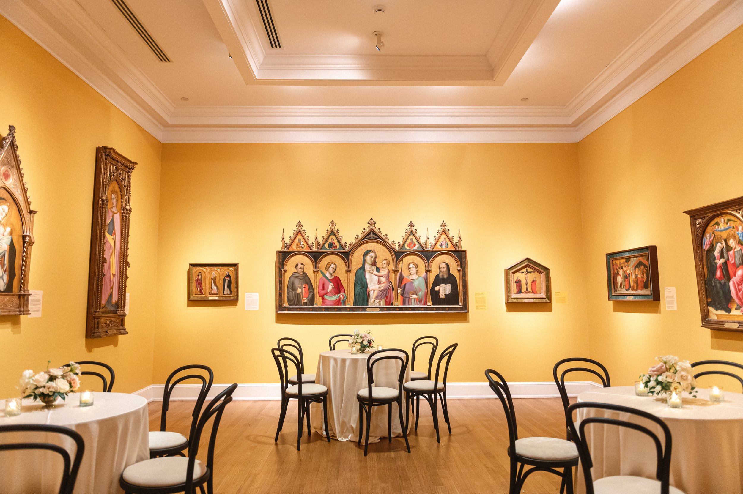 A room decorated for a celebration with round tables covered in white tablecloths, each with floral centerpieces and candles. Black chairs surround the tables. The yellow walls are adorned with religious paintings and a large multi-panel religious ic