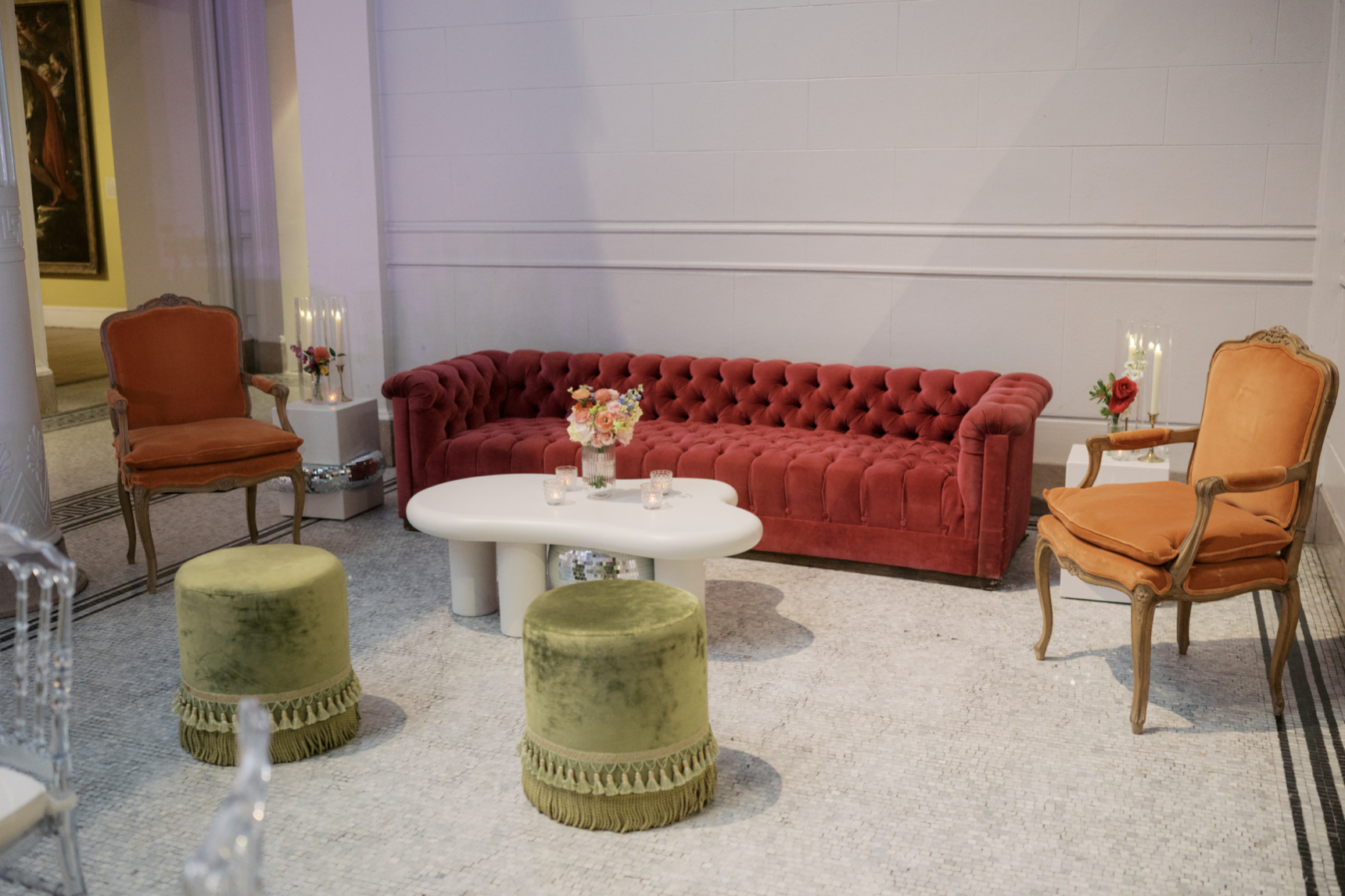 A vintage-style sitting area with a red velvet tufted sofa, two orange upholstered armchairs with wooden carved frames, two green velvet Ottoman stools, and white side tables with floral arrangements and candles, set on a terrazzo floor.