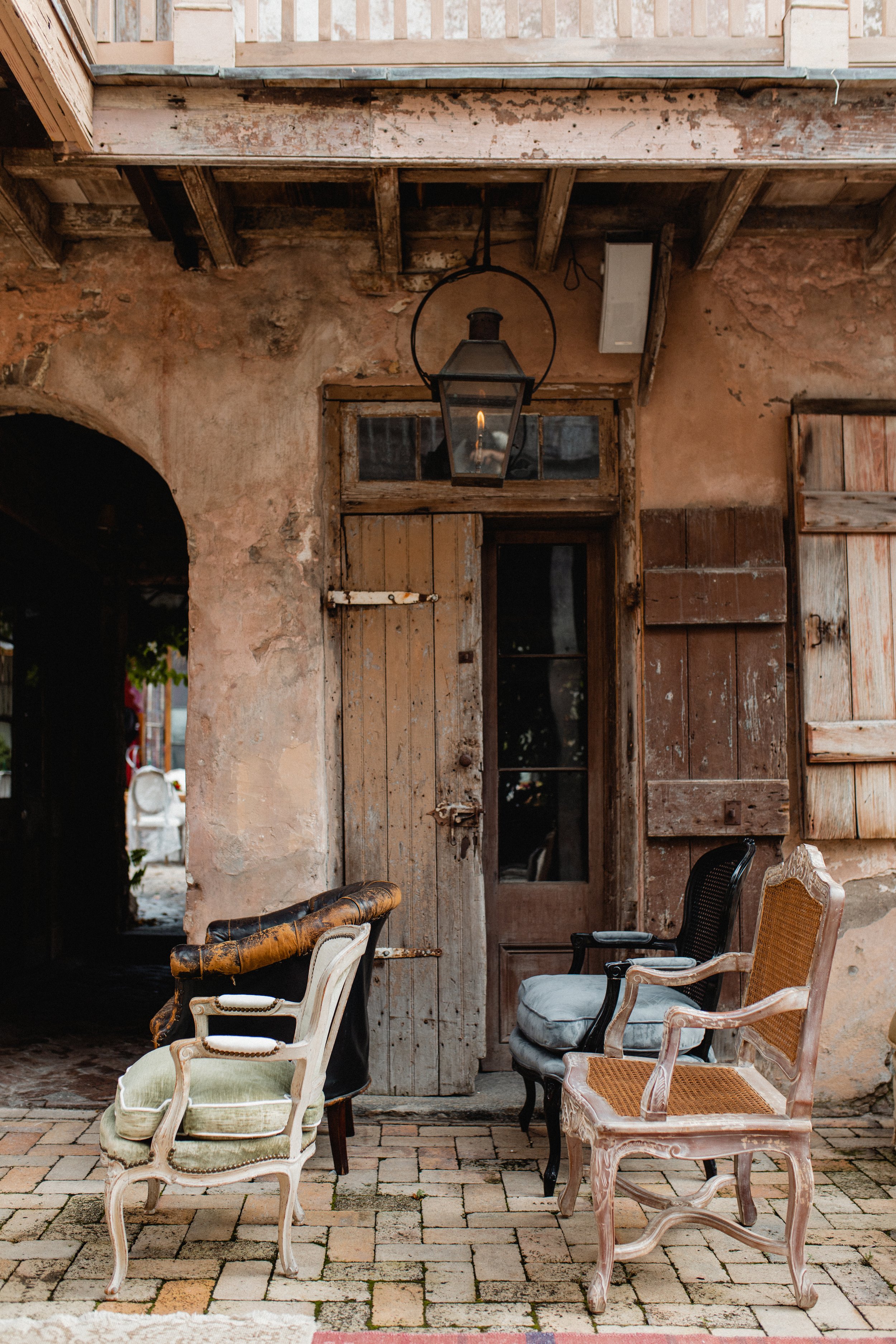 An outdoor covered patio with vintage chairs, a rustic door, and an old lantern hanging from the ceiling, featuring a weathered pink stucco wall and a brick floor.