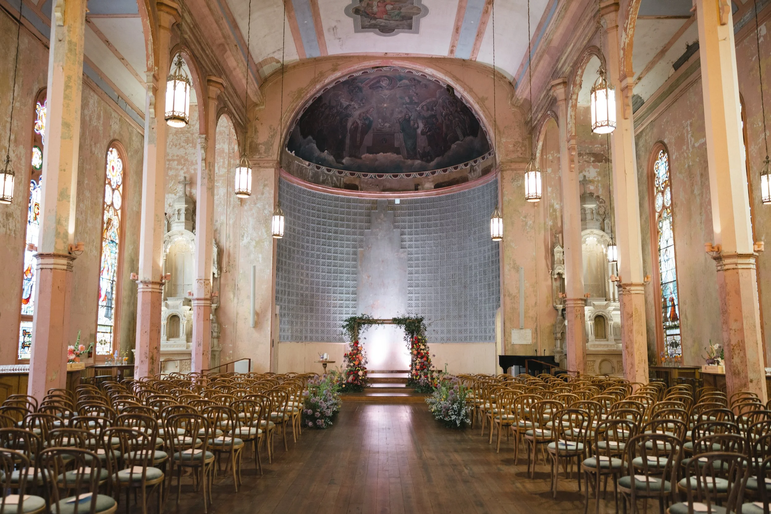 Interior of a church decorated for a wedding, with rows of chairs, floral arrangements, and a large religious mural on the ceiling.