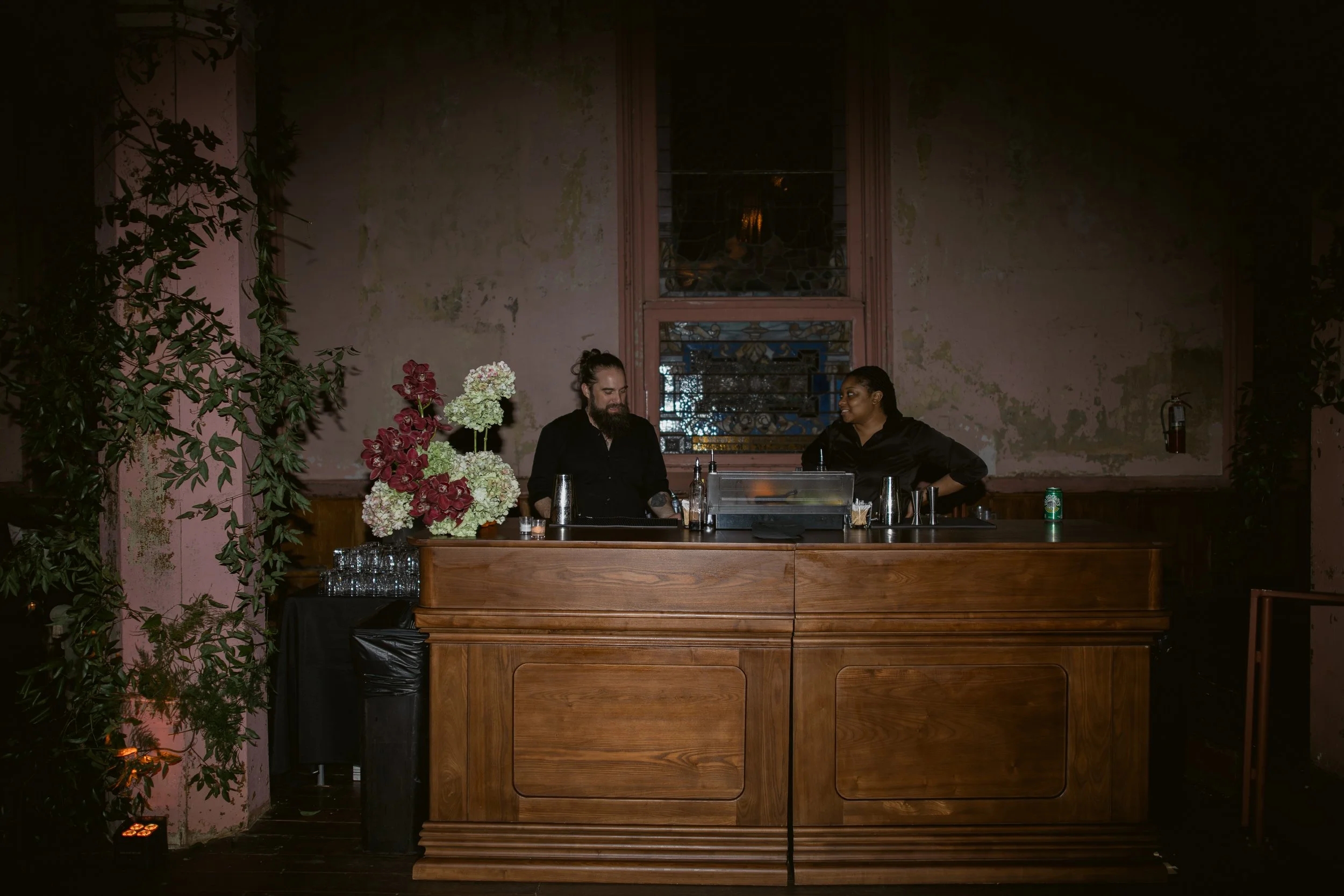 Two people standing behind a bar counter in a dimly lit, rustic room with weathered pink walls and a stained glass window. One is a man with a beard and a bun, and the other a woman with braided hair. There are flowers and bottles on the bar, with gl
