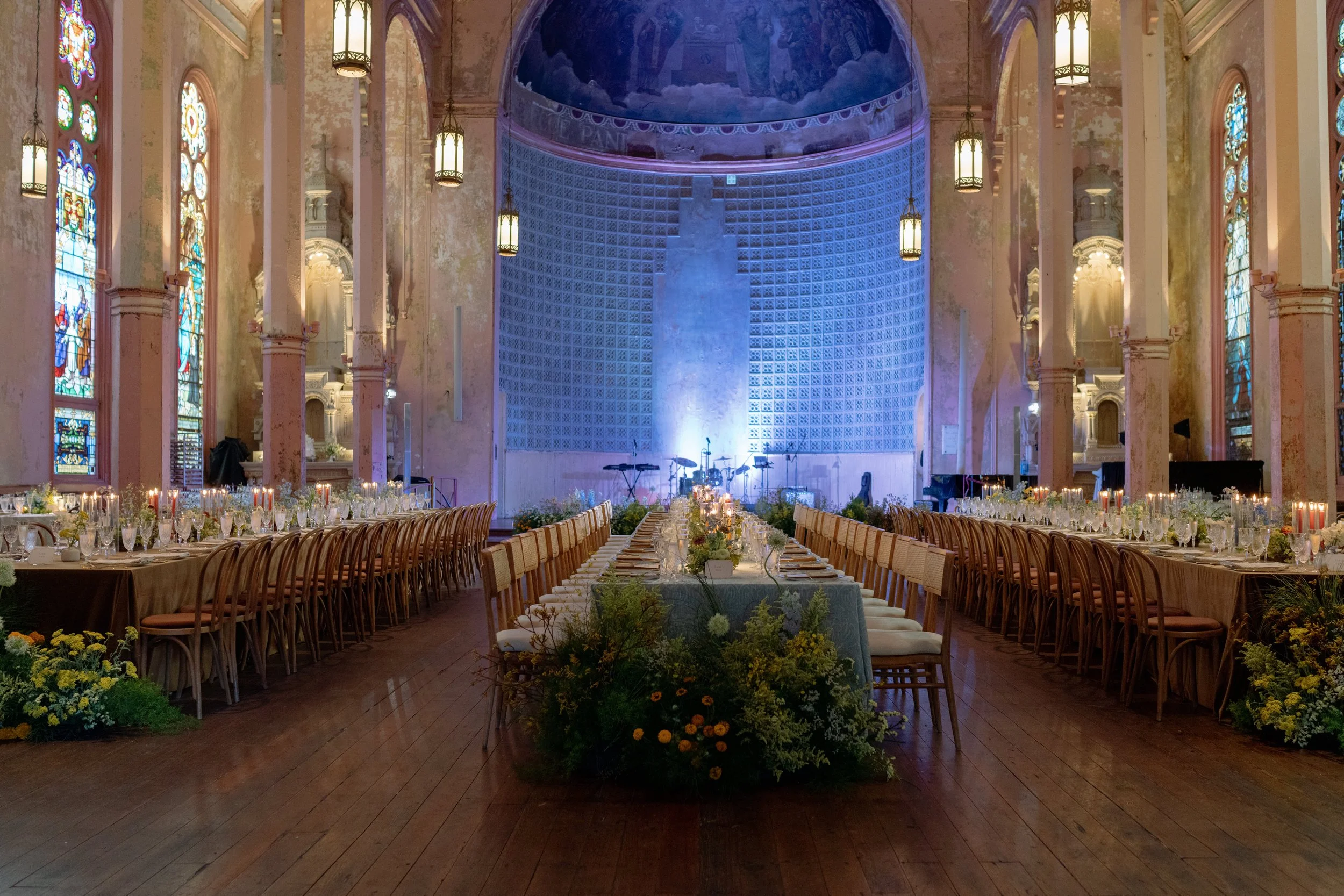 The image shows an elegant, large hall set up for a wedding or banquet with long tables arranged in the center, decorated with floral arrangements and candles. The hall features high ceilings, tall stained glass windows, and ornate chandeliers, with 