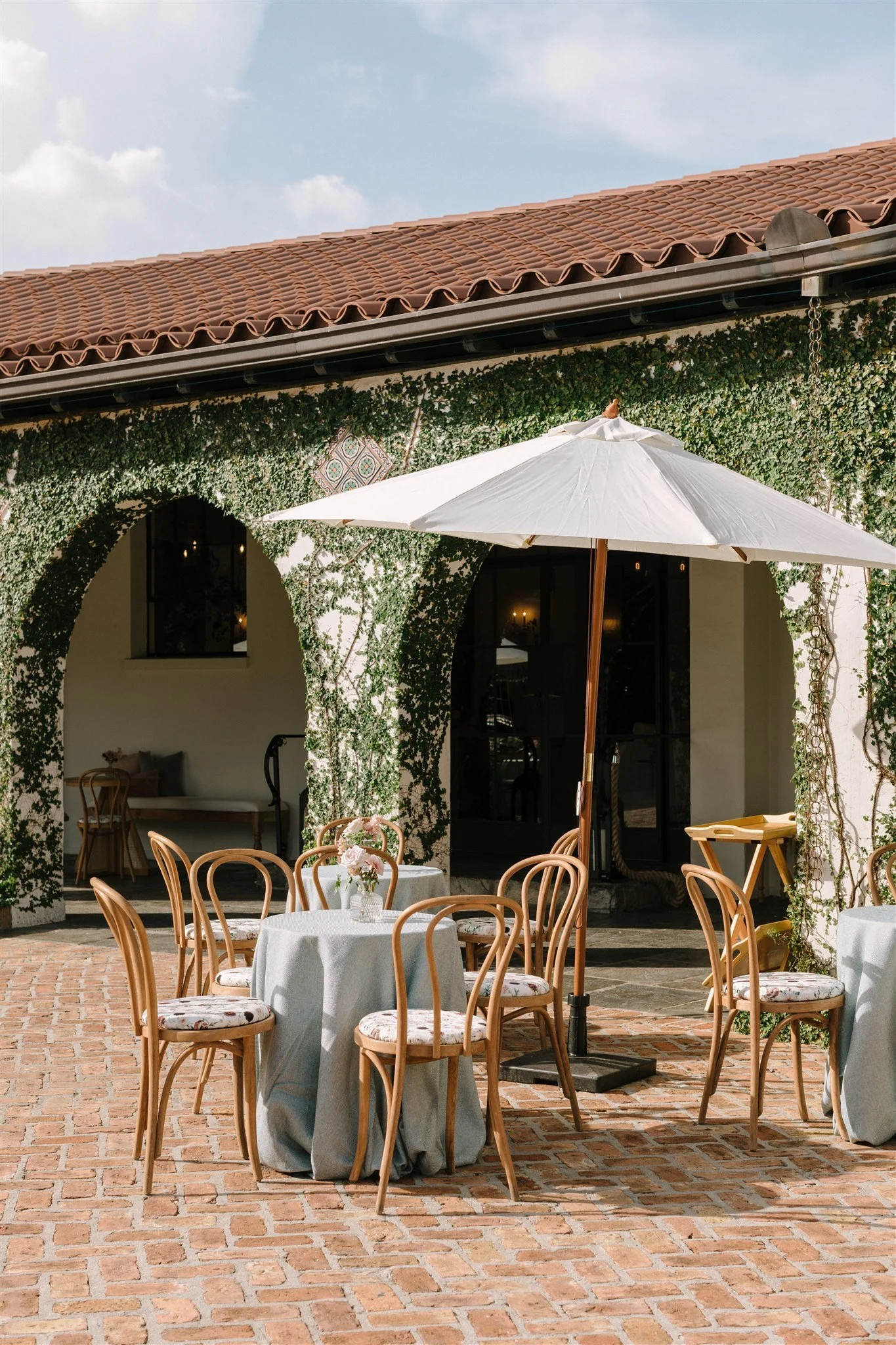 Outdoor patio area with round tables covered in white tablecloths, decorated with pink flowers in vases, surrounded by wooden chairs. There is a large white umbrella providing shade, and the background features a building with arched windows and a wa