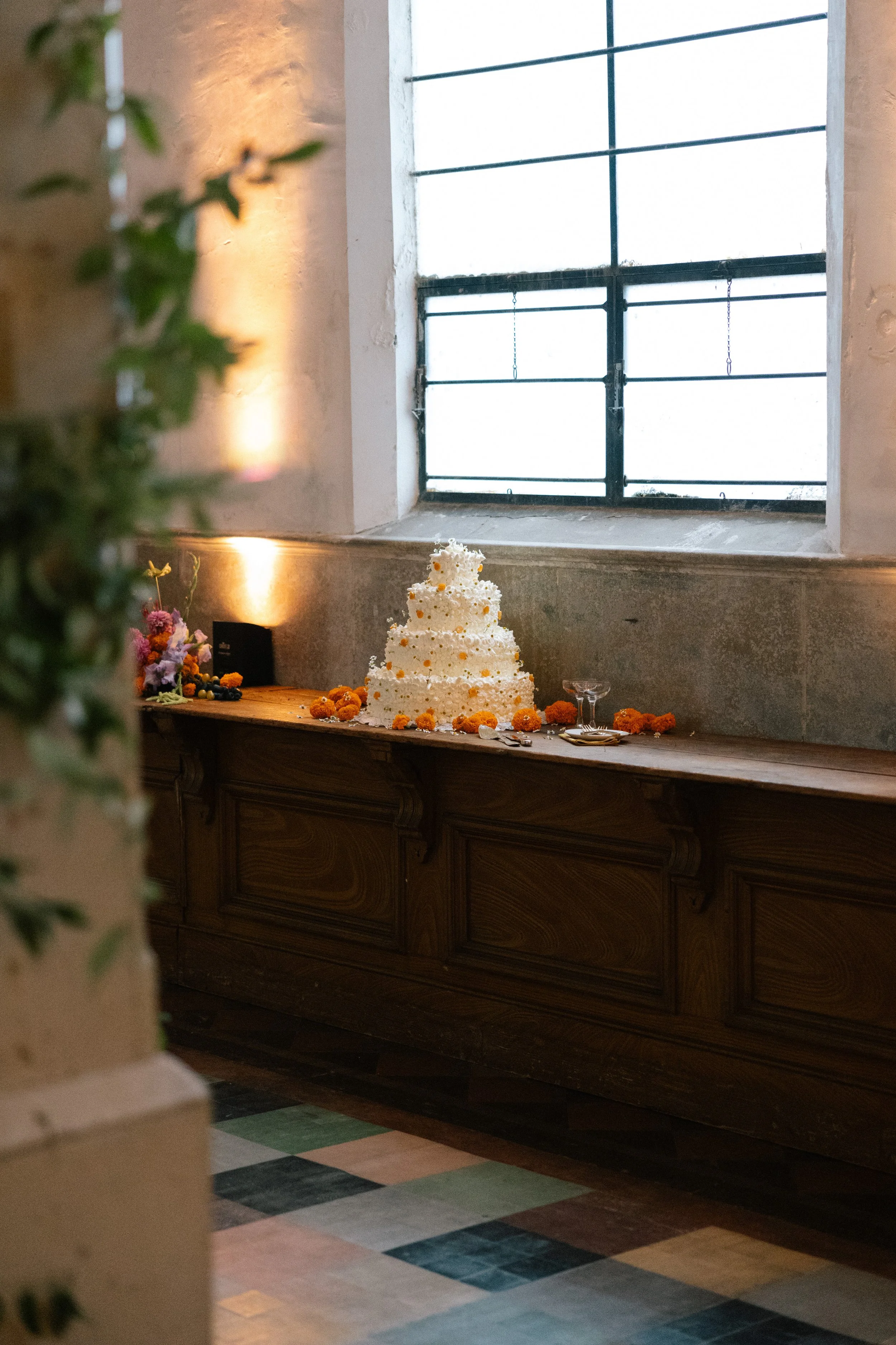 A decorative wedding cake with white frosting and orange floral accents sits on a wooden counter, surrounded by more flowers and tableware, in a rustic-style room with a large industrial window.