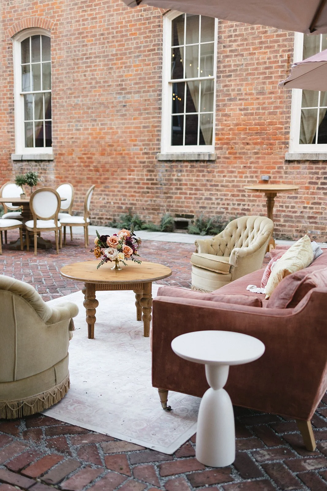 Outdoor patio with vintage furniture including a pink sofa, cream armchairs, a wooden coffee table with a floral arrangement, and a white side table, set against a brick wall with tall windows.