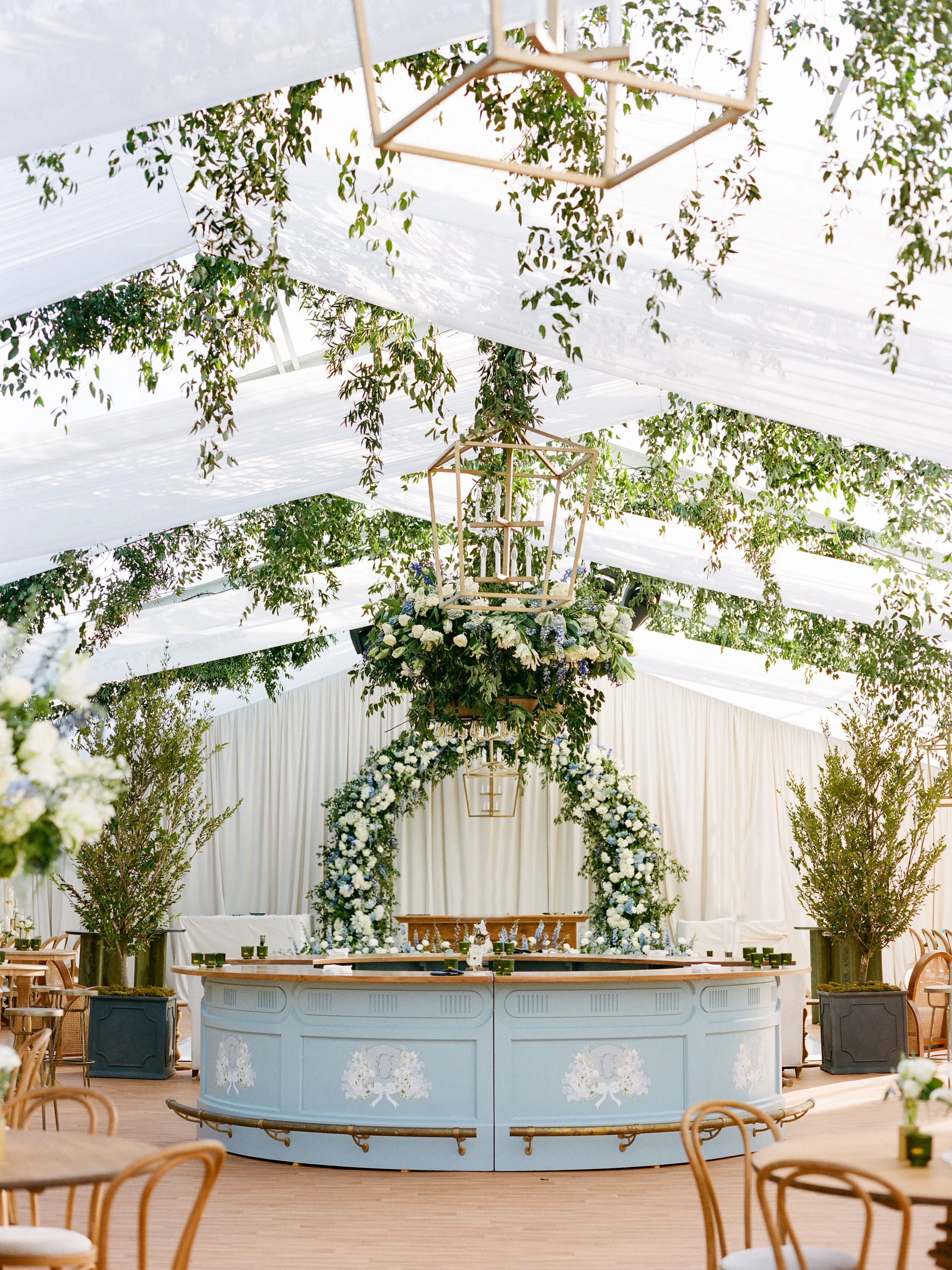 Interior of a wedding or event space decorated with flowers, greenery, and elegant lighting, featuring a curved bar in the foreground and a floral arch in the background under a white ceiling with hanging greenery.