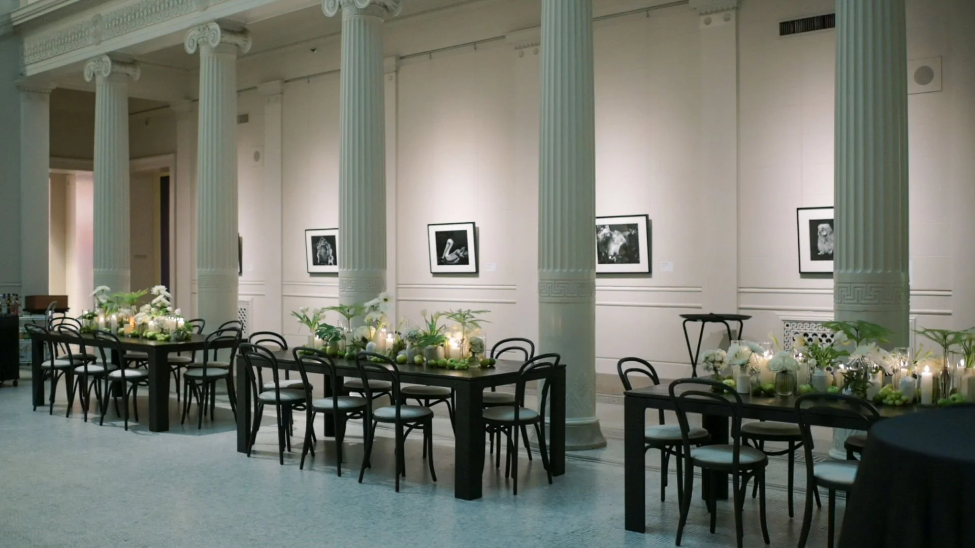Elegant event space with framed black-and-white photographs on the wall, decorated tables with floral arrangements, candles, green apples, and white flowers, surrounded by black chairs, in a room with tall white columns.