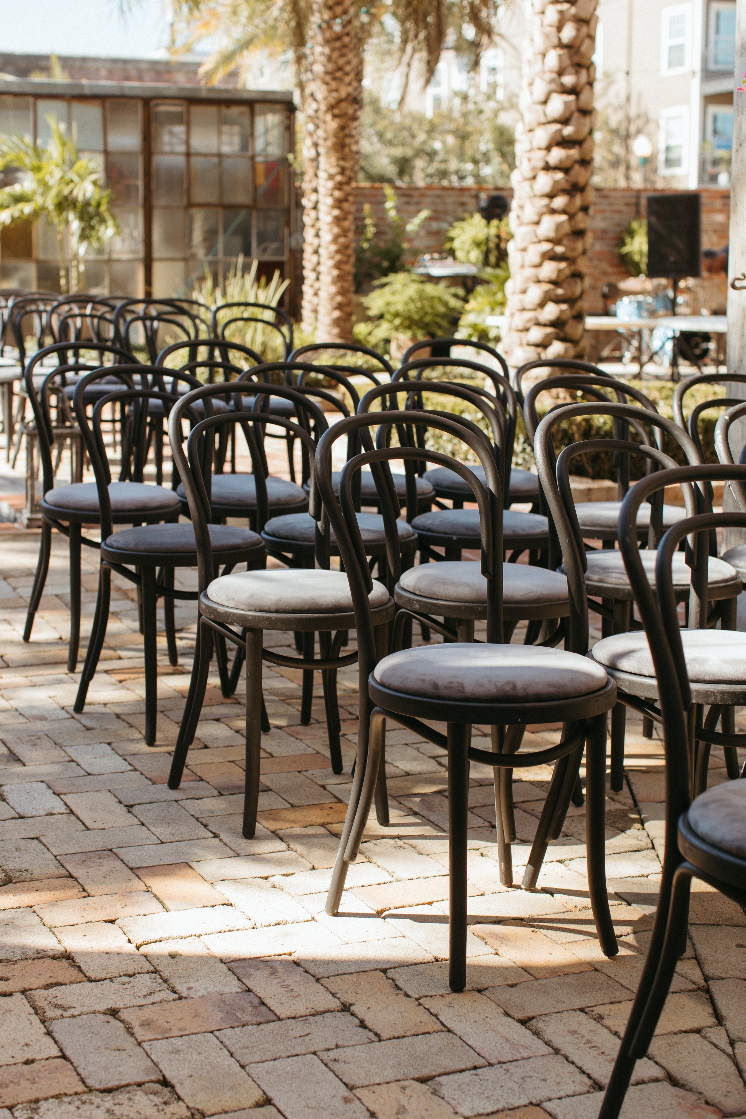 Outdoor seating area with multiple black chairs arranged on a brick patio, featuring palm trees, potted plants, and a brick wall in the background, with a DJ setup and some buildings behind.