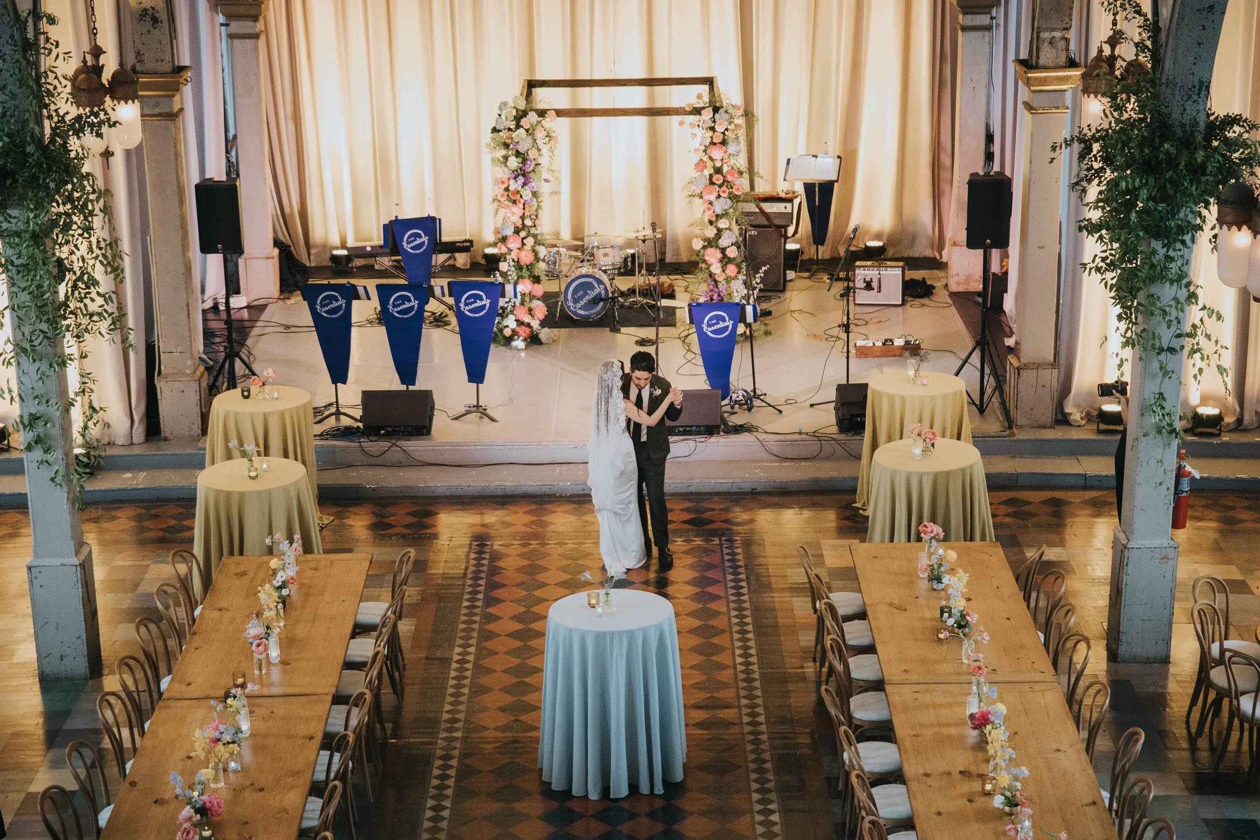 Wedding reception area with a couple dancing, decorated with pink and white flowers, tables with floral centerpieces, and a stage with musical instruments and floral arch.