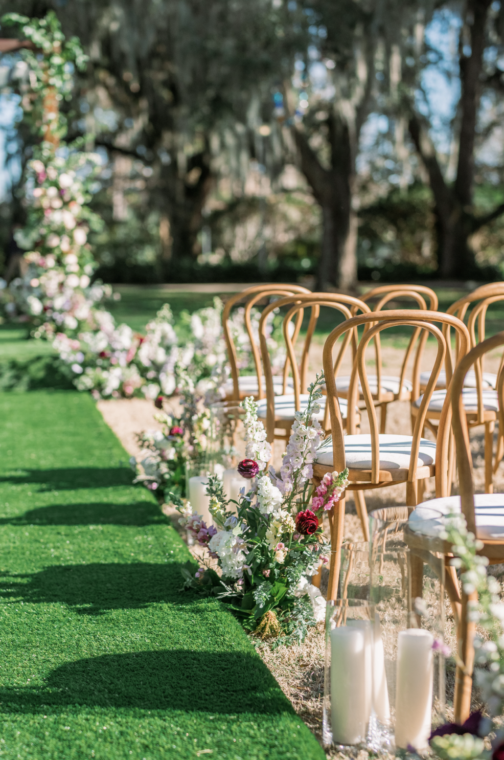 Outdoor wedding ceremony setup with wooden chairs arranged on a grassy aisle, decorated with white and pink flowers, candles, and trees in the background.