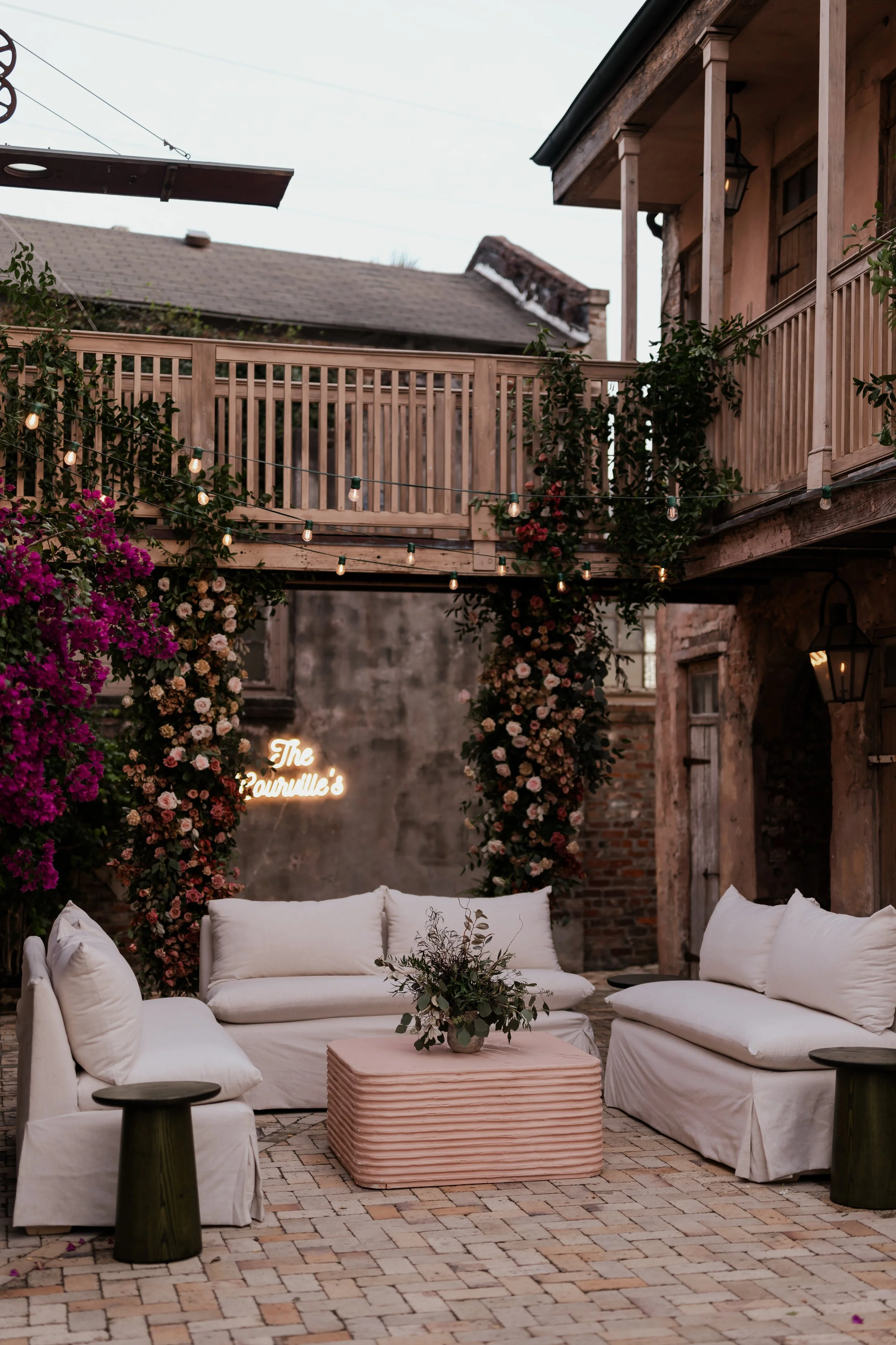 Outdoor patio seating area with white sofas, a pink textured coffee table, small black side tables, string lights, and floral decorations in a rustic setting.
