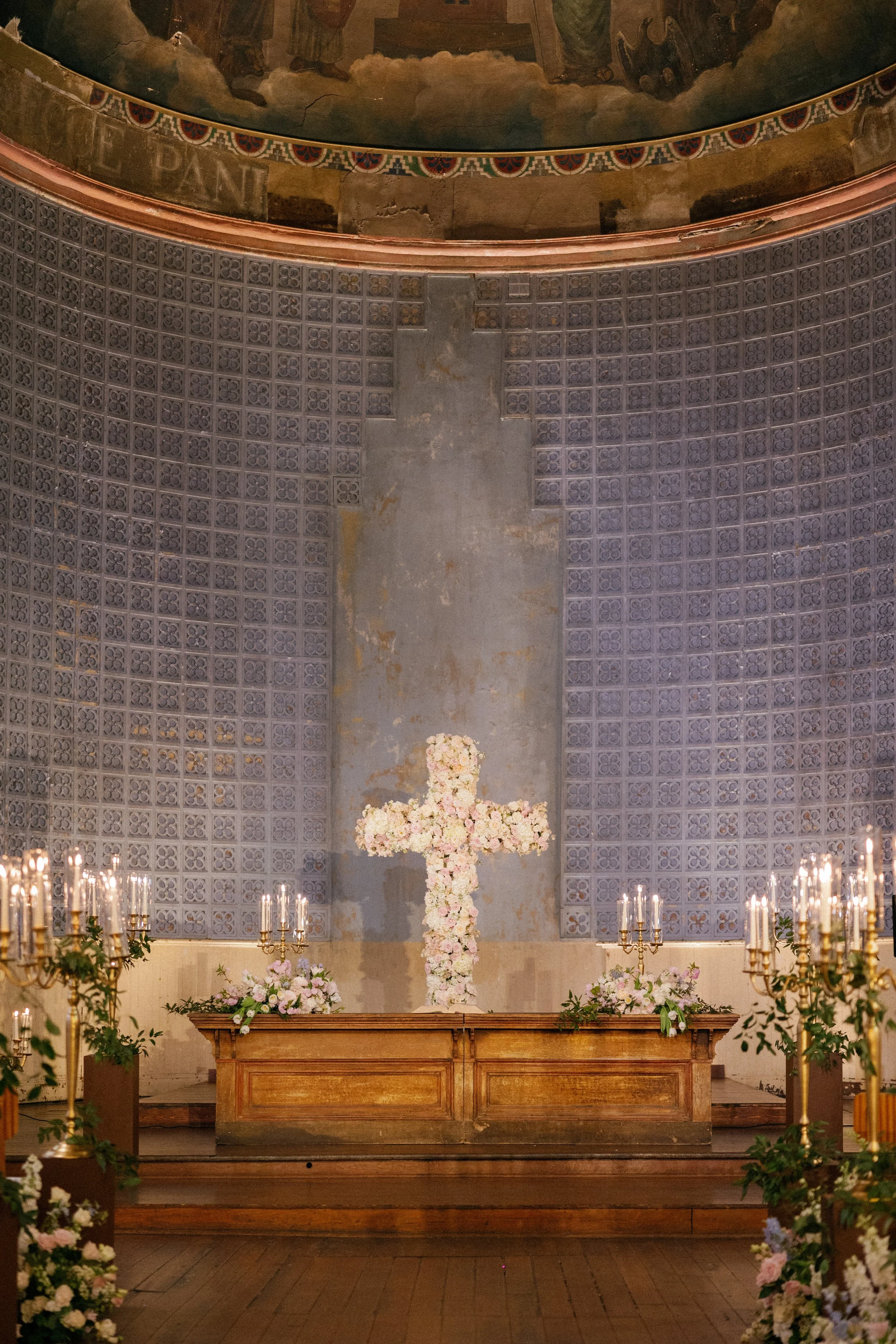 Interior of a church or chapel decorated for a wedding, featuring a large cross made of pink and white flowers at the altar, surrounded by floral arrangements and candles.