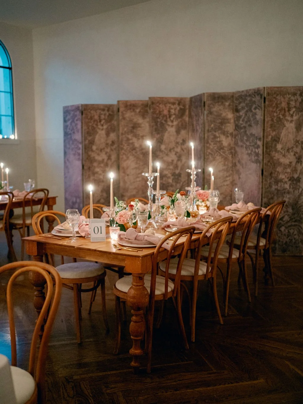 Elegant wedding reception table with pink floral centerpiece, tall candle holders with lit candles, wine glasses, napkins, and table setting, in a softly lit room with a decorative foldable screen and arched window.