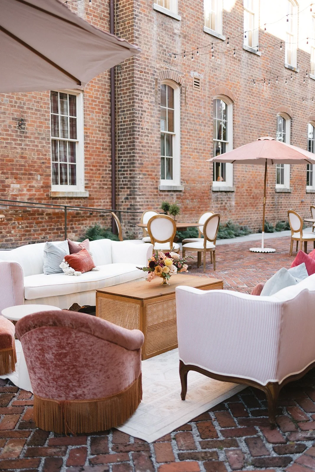 Outdoor patio area with vintage-style furniture, including a white sofa with pillows, pink velvet armchairs, a wooden coffee table with a flower arrangement, round dining chairs, and large beige umbrellas. The background features a brick building wit