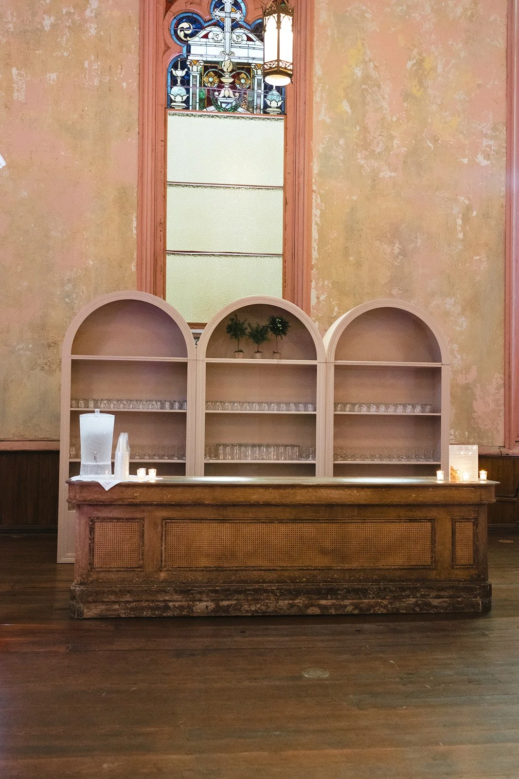 Empty vintage bar with three arched shelves behind it, a stained glass window with a cross at the top, and a hanging lantern light, with candles and a water dispenser on the bar.
