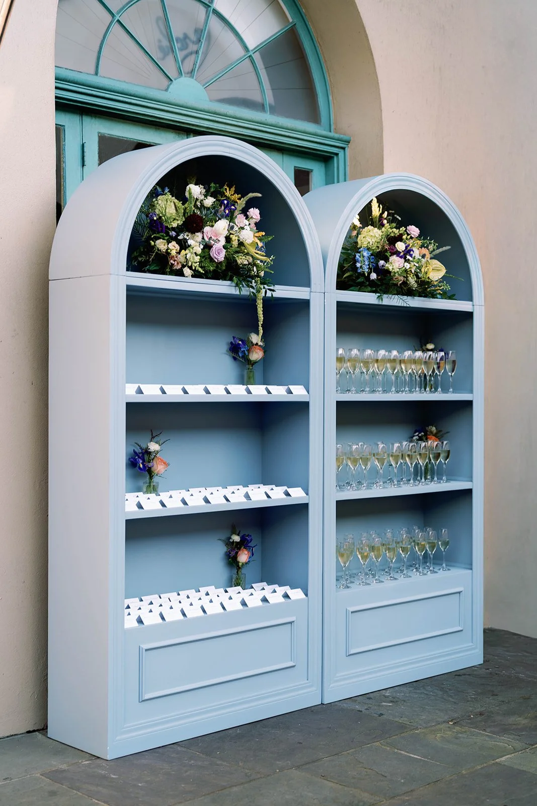 White display cabinet with floral arrangements and champagne glasses against a beige wall outside a building.